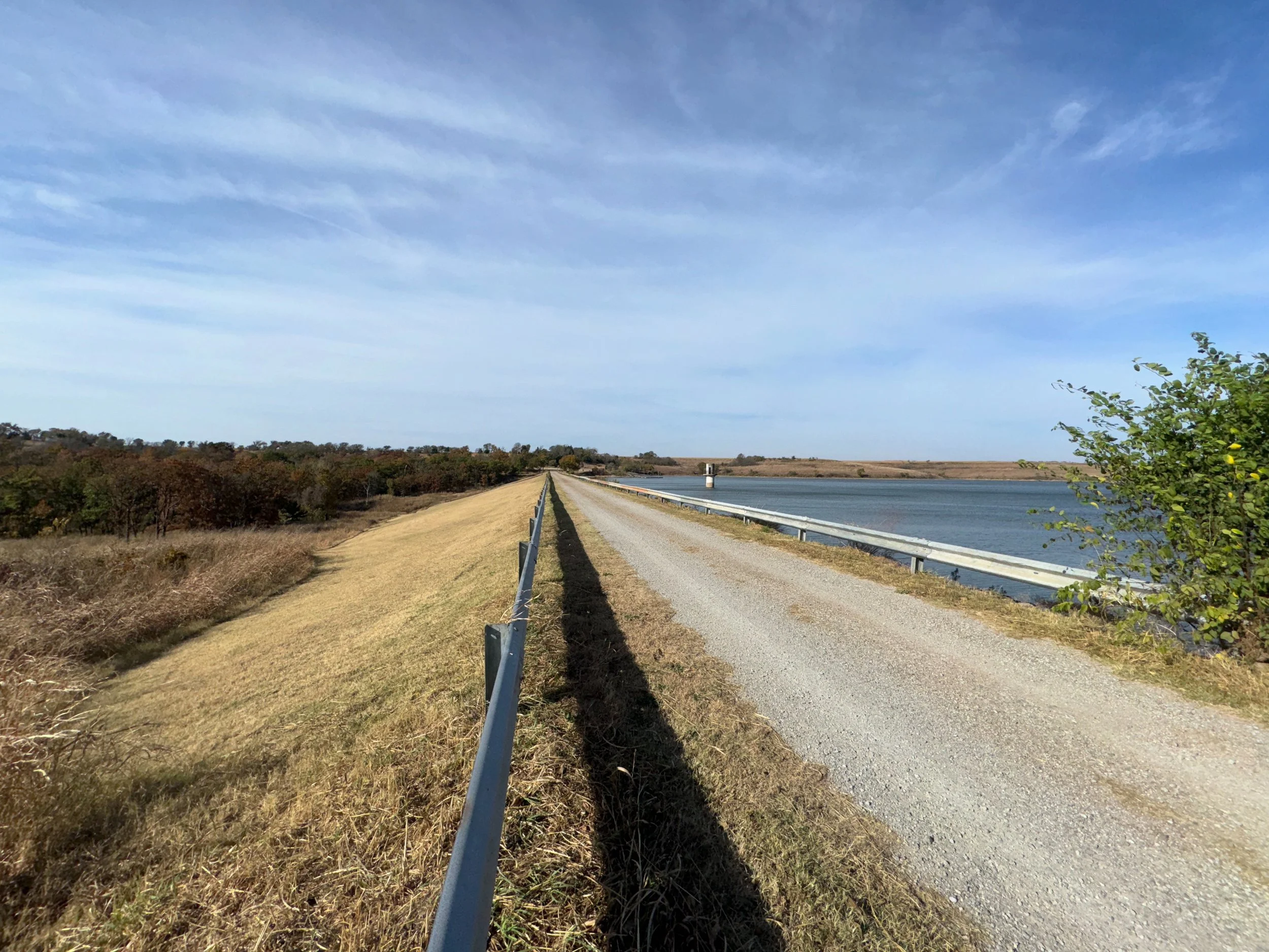 Rural dirt road beside a body of water and grassy field under blue sky