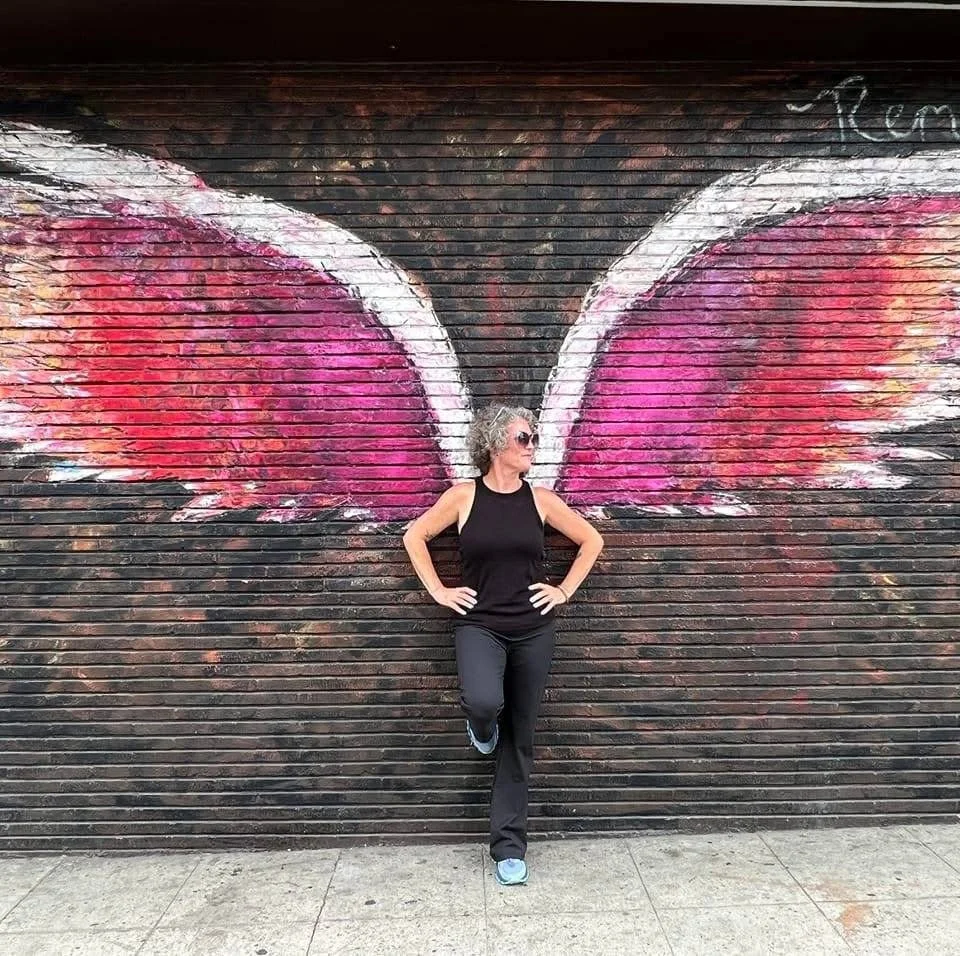 Woman with curly hair and sunglasses standing in front of a brick wall painted with pink and white butterfly wings.