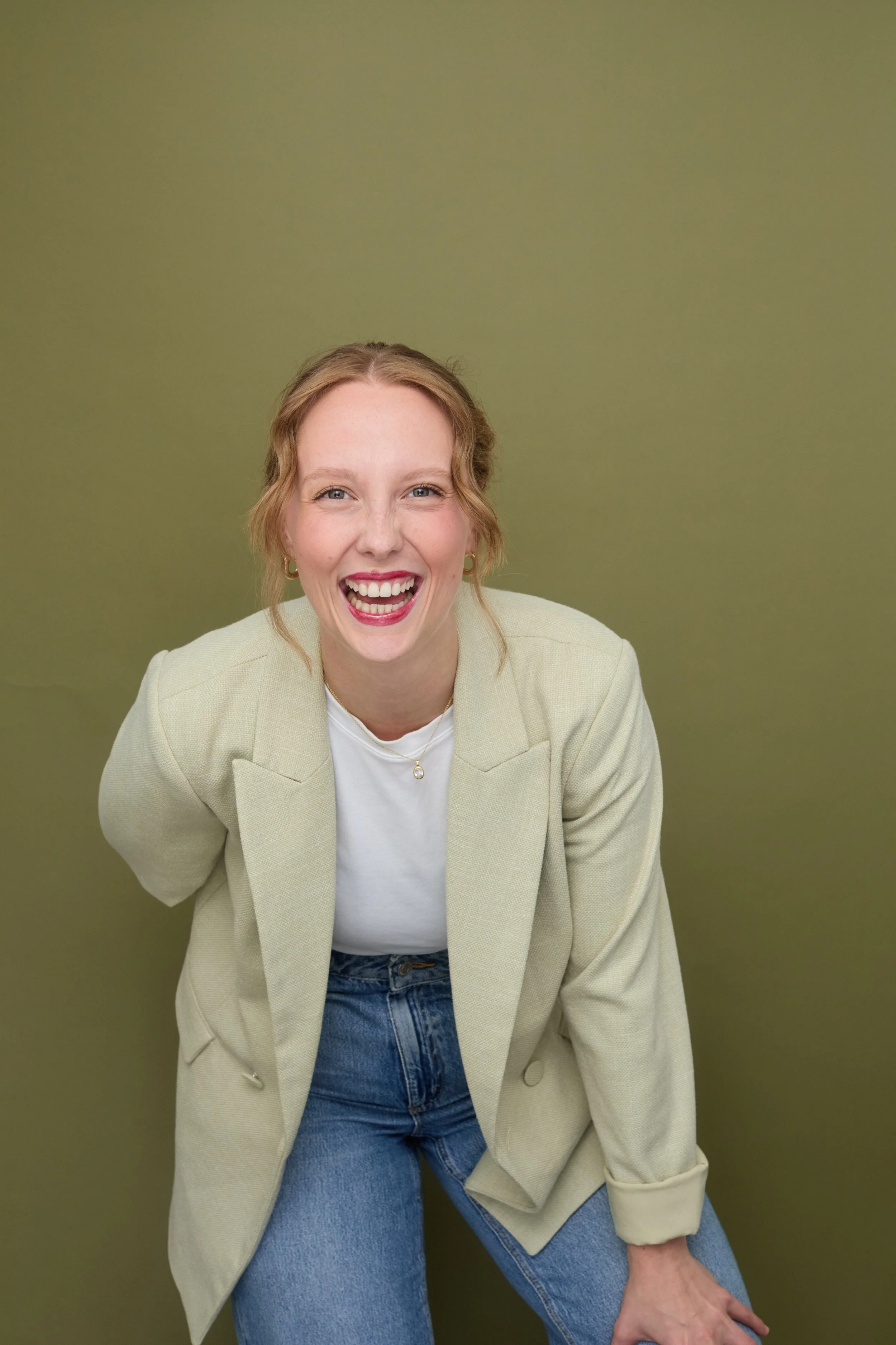A woman with red hair and a big smile, wearing a beige blazer, white top, and blue jeans, posing against a green background. Rachel Anne Hunt
