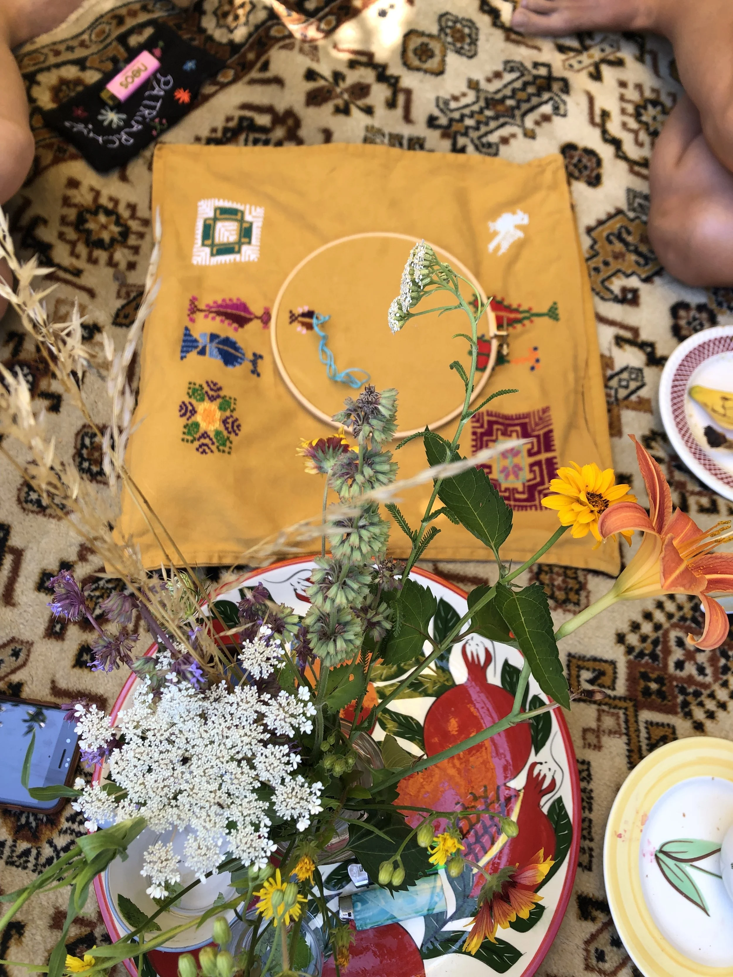 A floral arrangement with colorful flowers placed on a table with a patterned tablecloth. Behind is a yellow cloth with embroidered patterns, a bracelet, and some other small items. To the side, there are plates, a smartphone, and part of a person's 