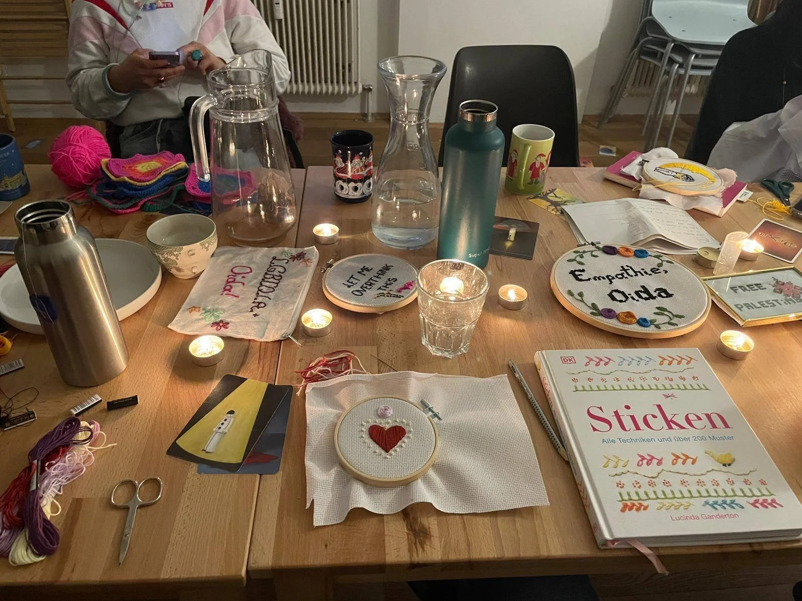 A table with embroidery projects, threads, embroidery hoops, candles, water bottles, mugs, and a person with knitting yarn in the background.