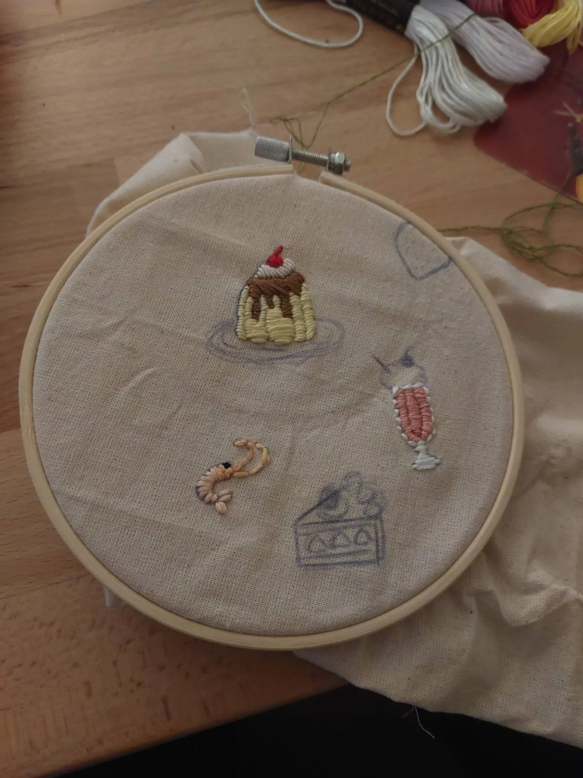 Embroidery hoop with partially completed embroidered designs of a beer mug, a cocktail, a shrimp, and a cake on fabric.