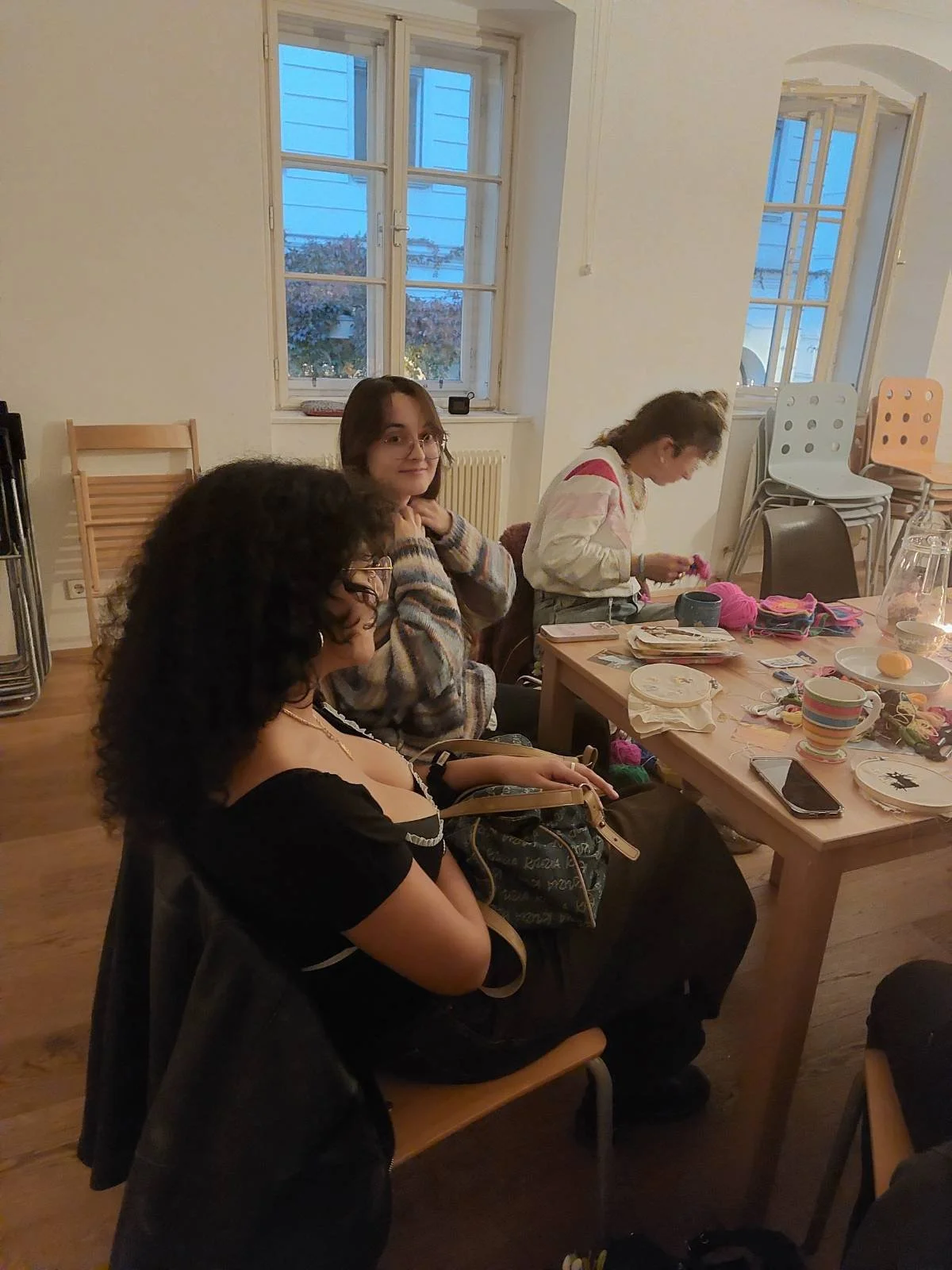 Three women sit at a wooden table in a cozy room with large windows showing a blueish evening sky. The table has various craft supplies, cups, and a smartphone. The women are engaged in knitting or crocheting activities.