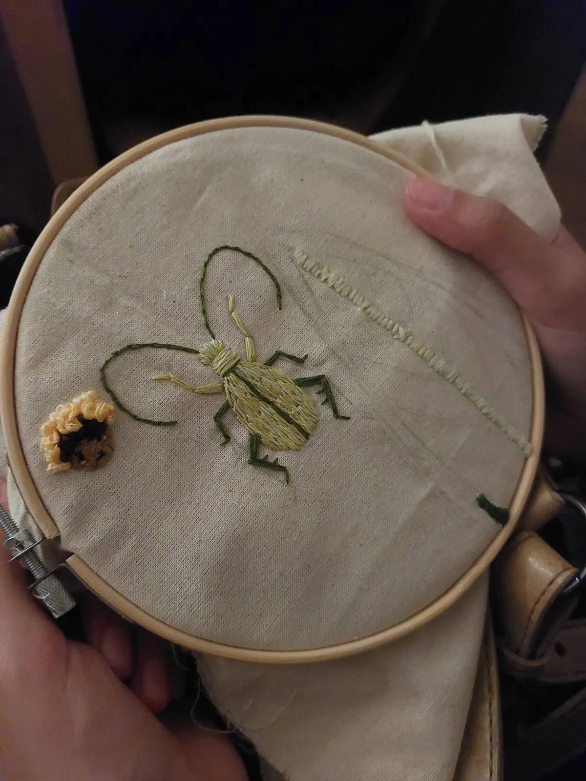 Close-up of embroidery in a hoop, featuring a bee with green and yellow threads, a small round flower, and a partially stitched leaf.