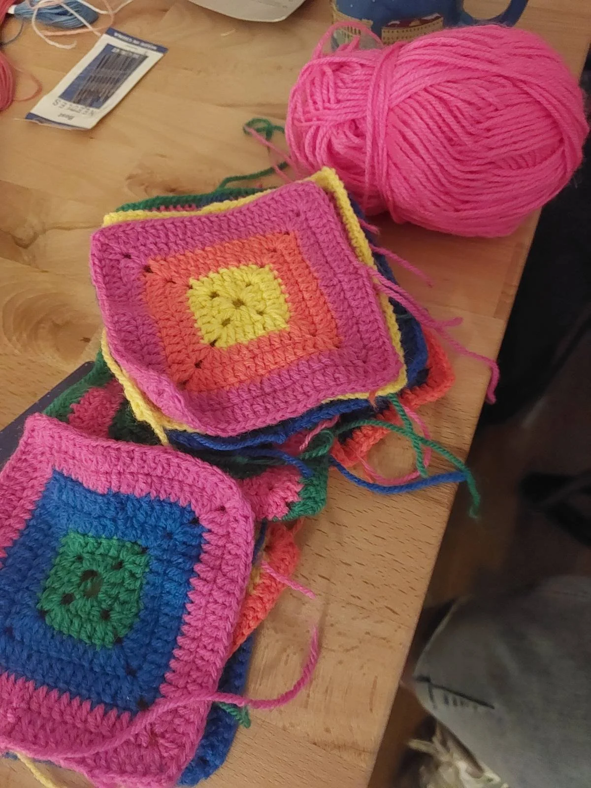 Colorful crocheted granny squares in pink, yellow, orange, blue, green, and black yarn on a wooden table, with a ball of pink yarn nearby.
