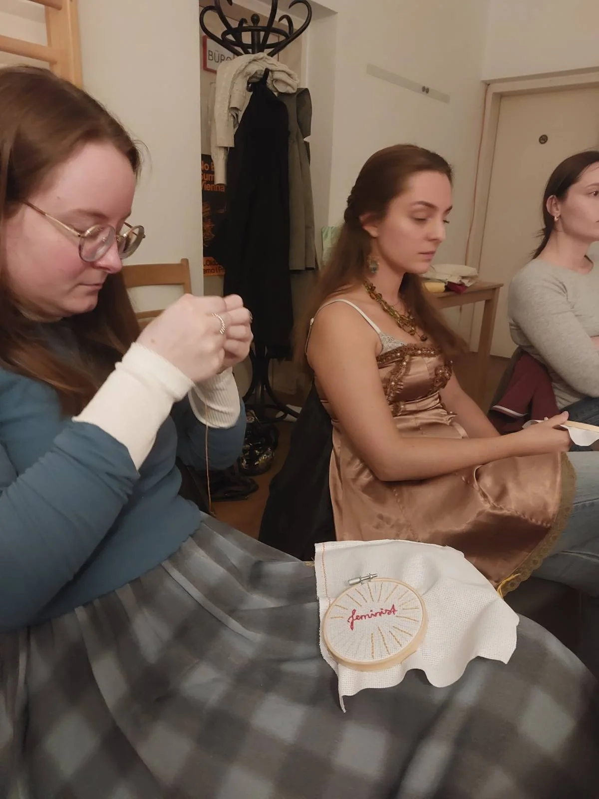 A woman with glasses knitting with a needle, with a piece of fabric and embroidery hoop on her lap, in a room with three women sitting at a table, one in a satin dress, another in a gray shirt, and a coat hanging in the background.