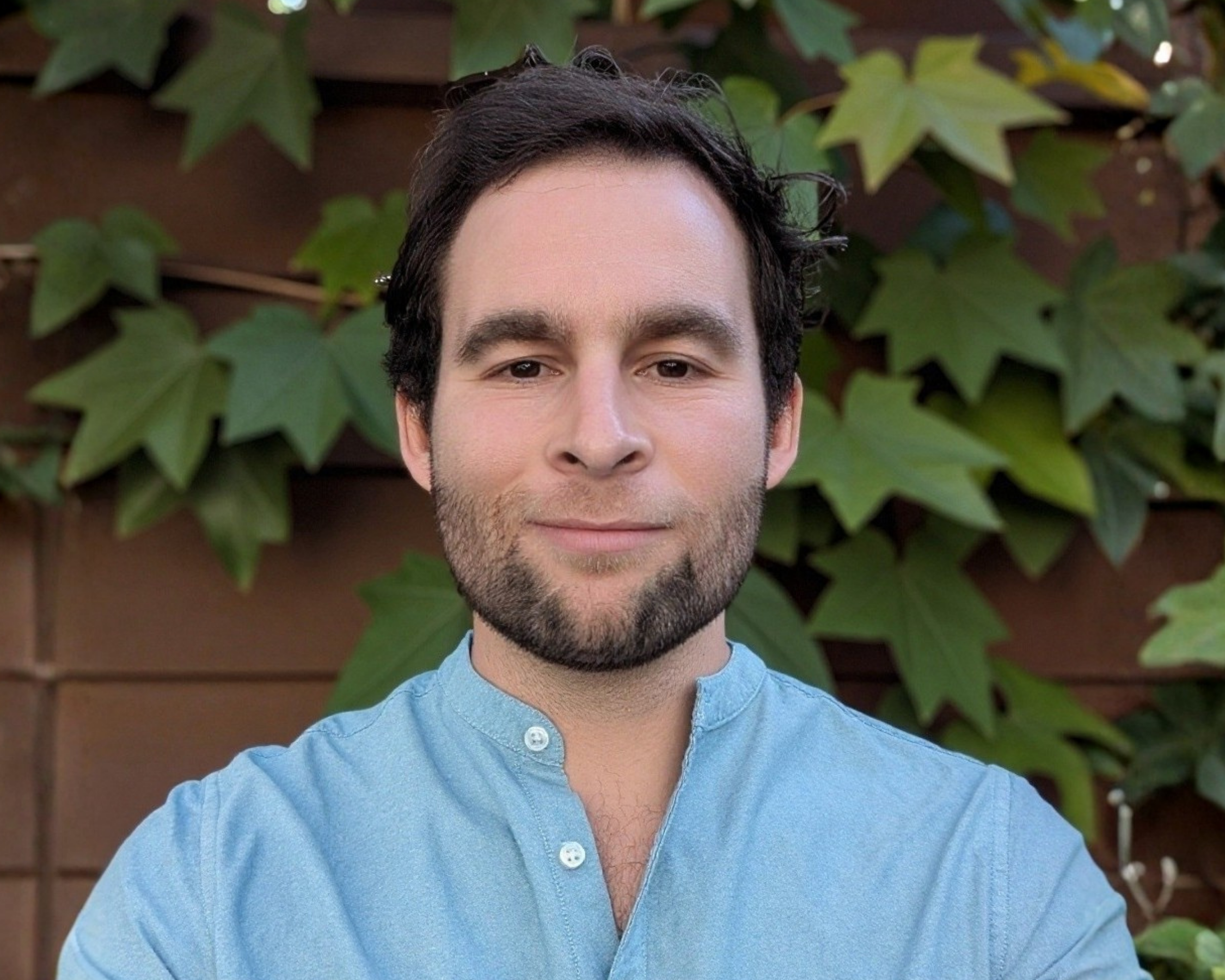 Head-and-shoulders portrait of a bearded man in a light blue shirt standing in front of leafy greenery.