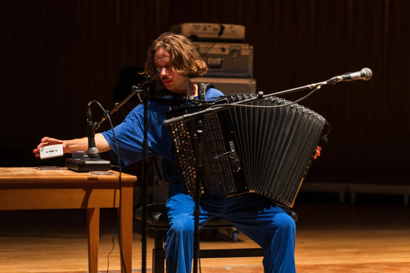 A man in blue clothing plays an accordion while sitting on a chair on stage, with microphones and equipment around him.