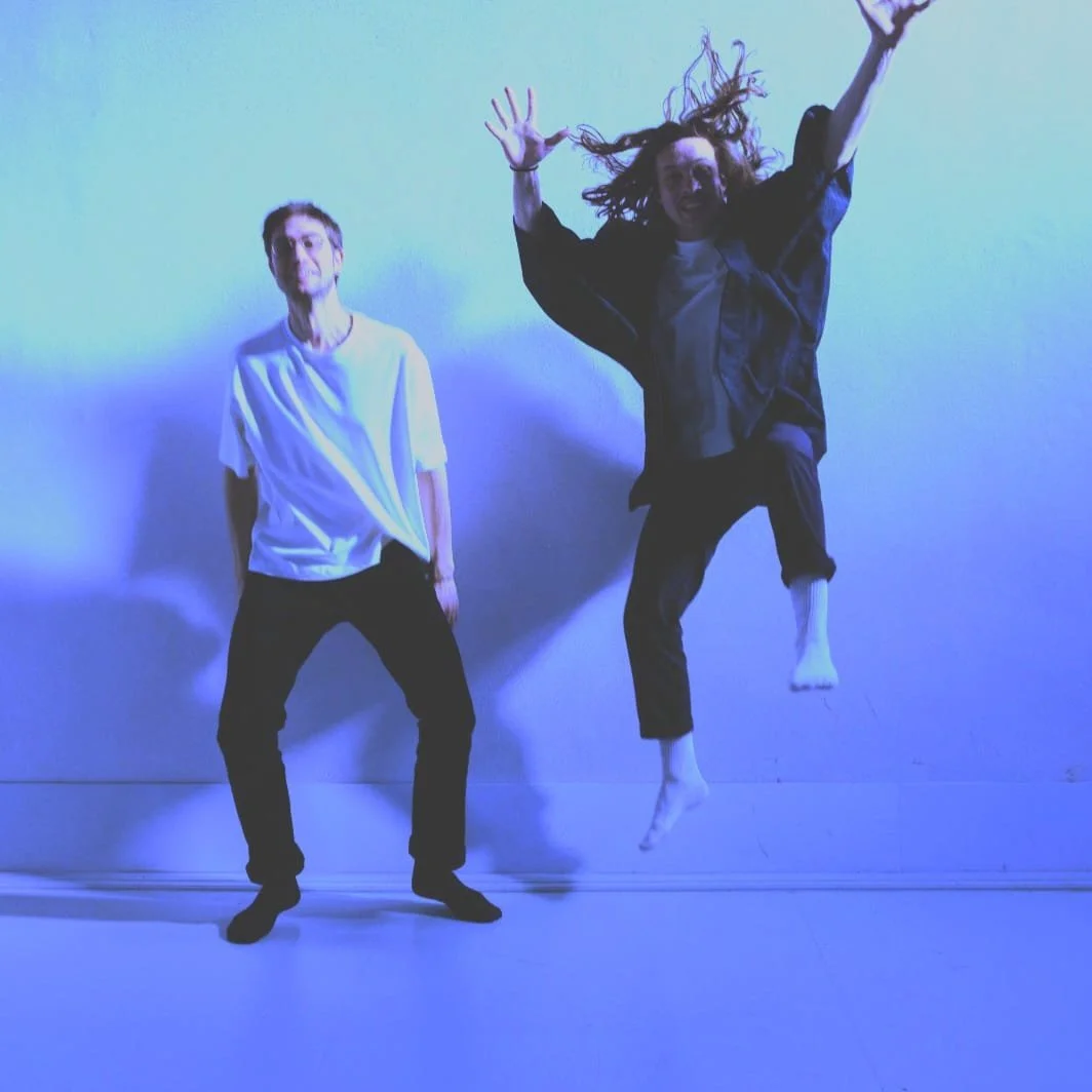 Two people, one standing and one jumping in the air, pose against a plain wall with blue lighting.