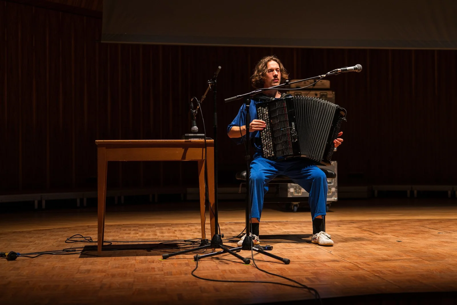 A man in blue clothing playing the piece Self Care by Jennifer Walshe on an accordion on stage with a microphone and a wooden table.