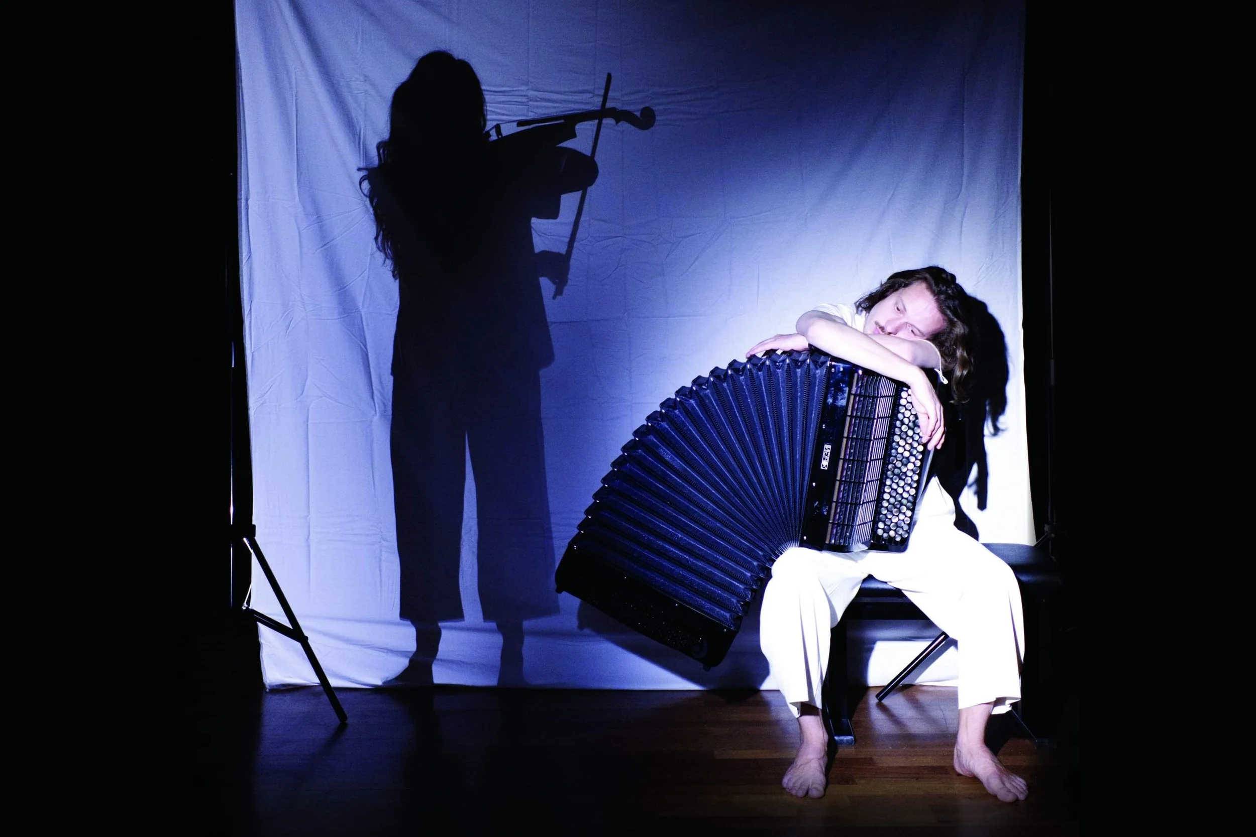 A man sitting on a chair, hugging an accordion, with his eyes closed and a peaceful expression, while a shadow of a woman playing the violin is cast on a white backdrop behind him.