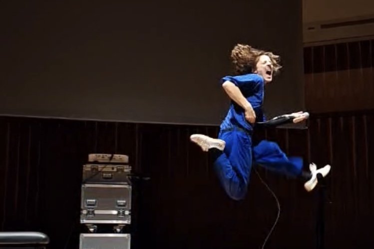 Person in blue scrubs jumping on stage while performing Self Care by Jennifer Walshe while holding a microphone with a joyful expression, dark background, some audio equipment visible.