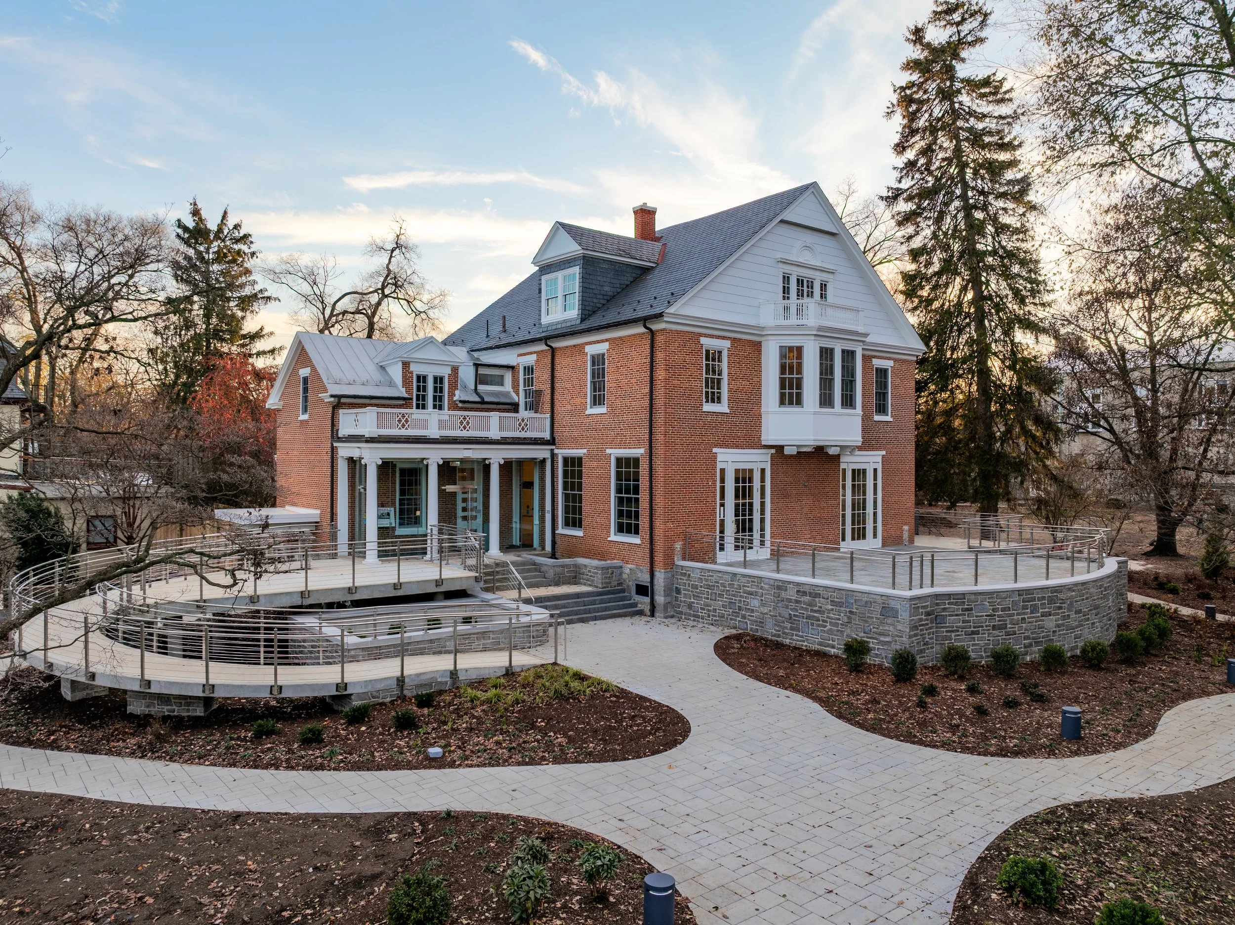 A large brick and white house with a gabled roof and multiple windows, surrounded by trees and an outdoor walkway with a curved pattern.