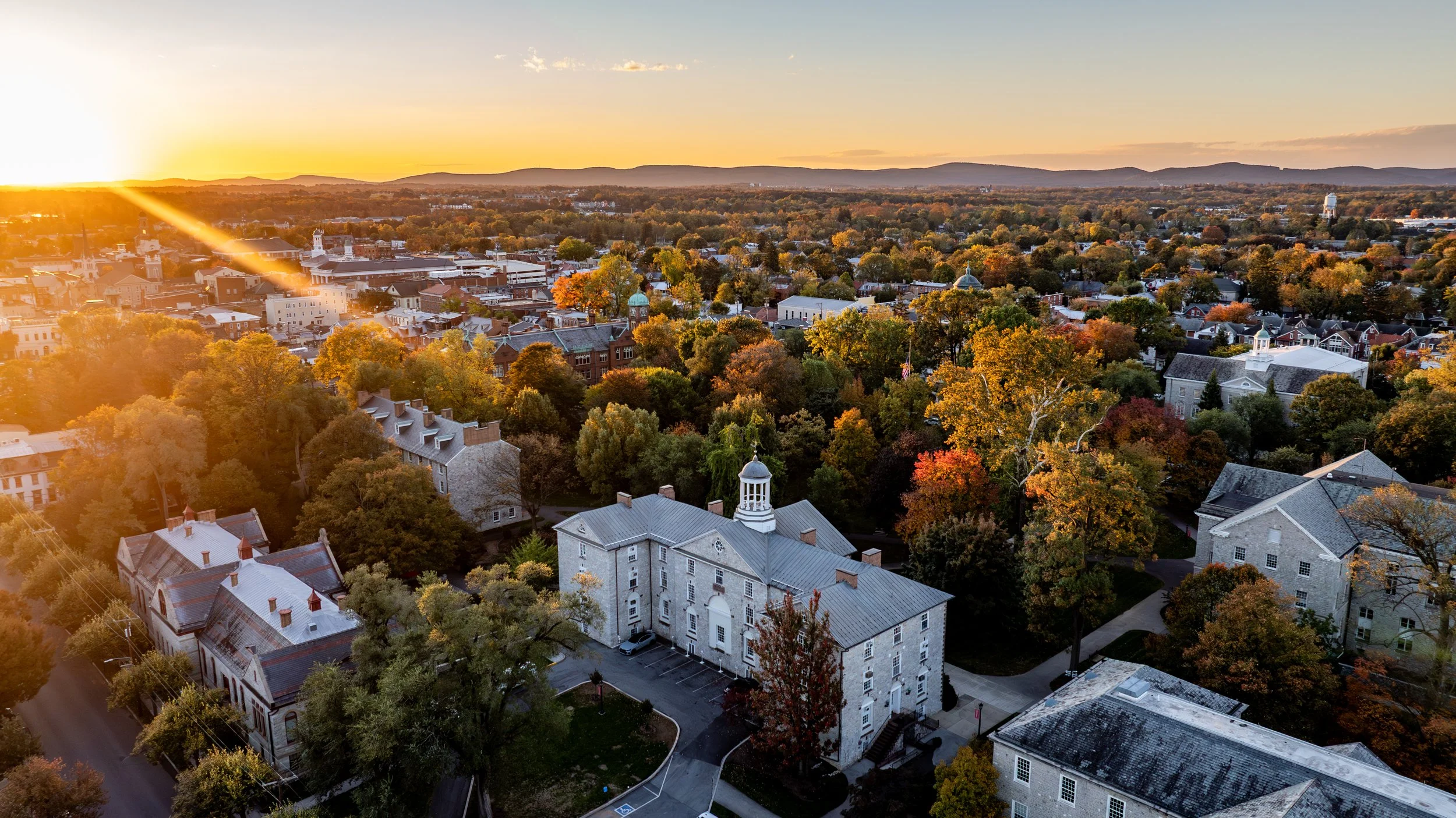 An aerial view of a small town during sunset, with trees displaying fall colors, historic buildings, and rolling hills in the background.