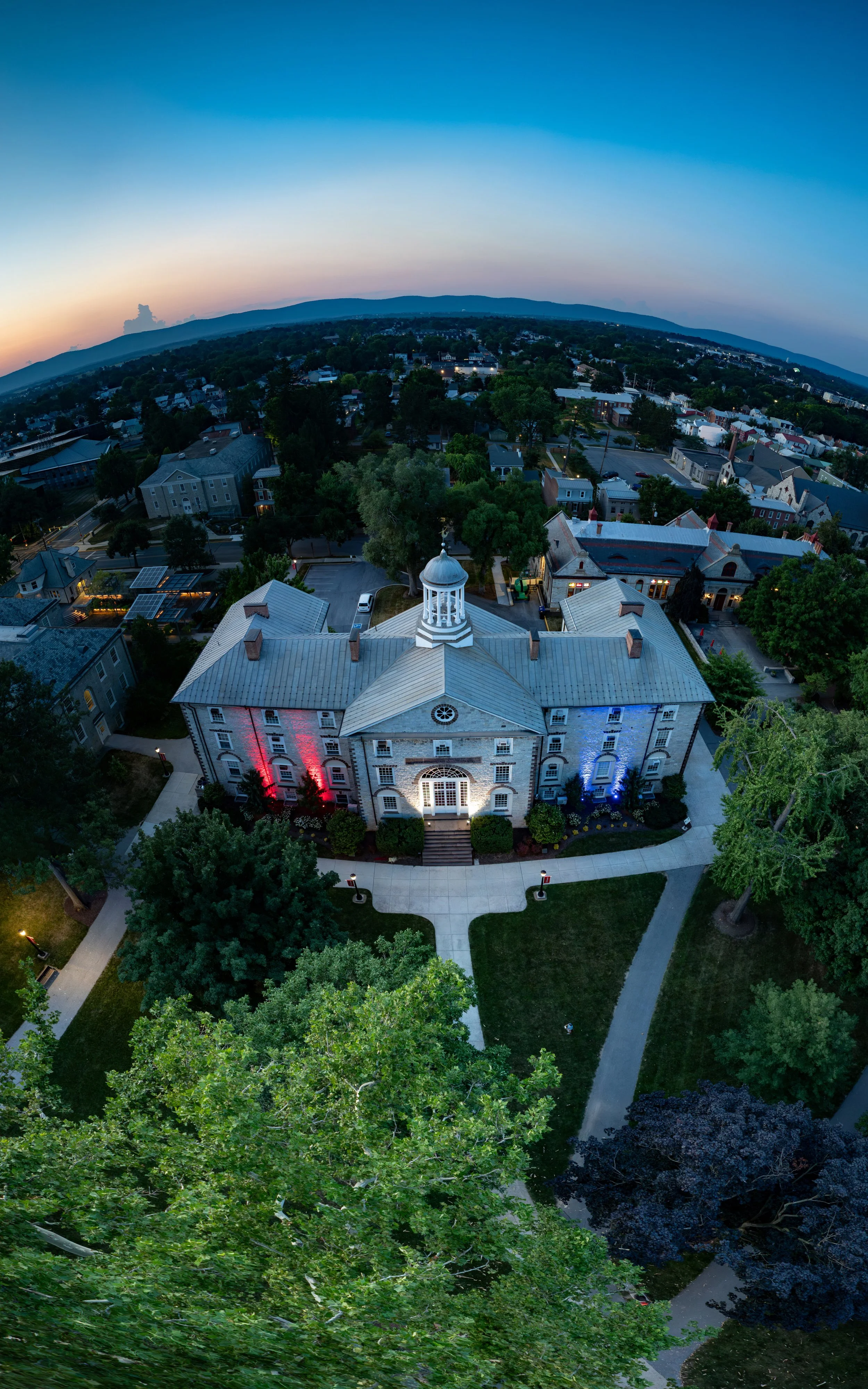 Aerial view of a historic building with a white cupola, surrounded by green trees and a small park, during dusk.