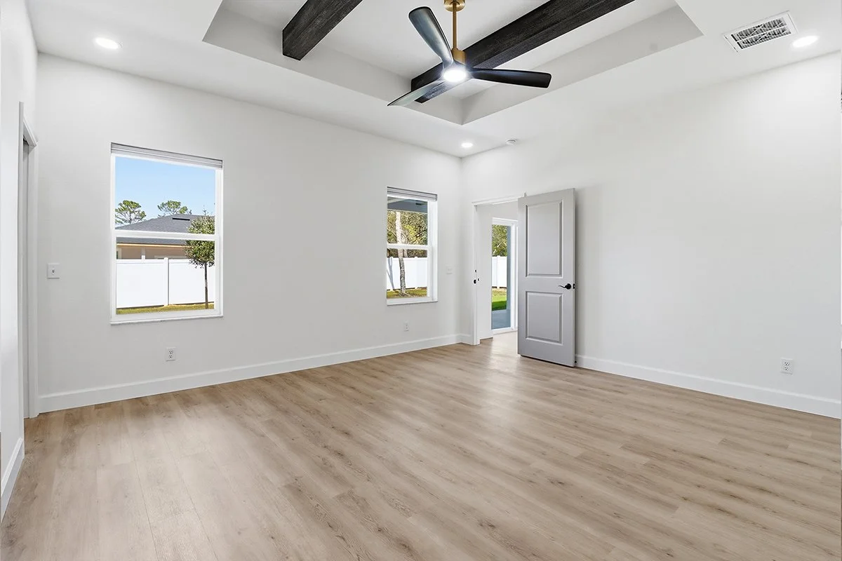 Primary bedroom in a Citrus Springs custom home featuring a tiered tray ceiling with dark wood beams and large windows overlooking the backyard.