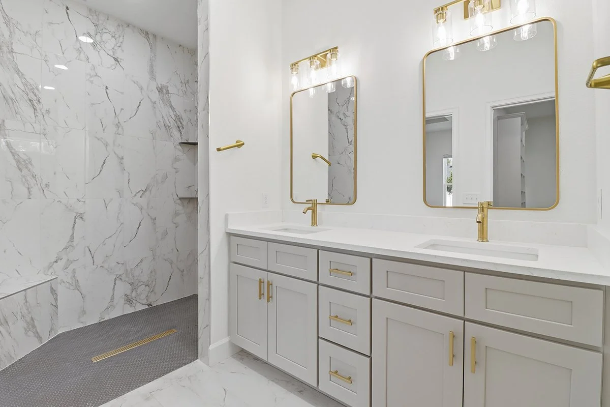 Modern bathroom with gray shaker cabinets, white quartz counters, gold-framed mirrors, and a floor-to-ceiling marble-tiled shower.