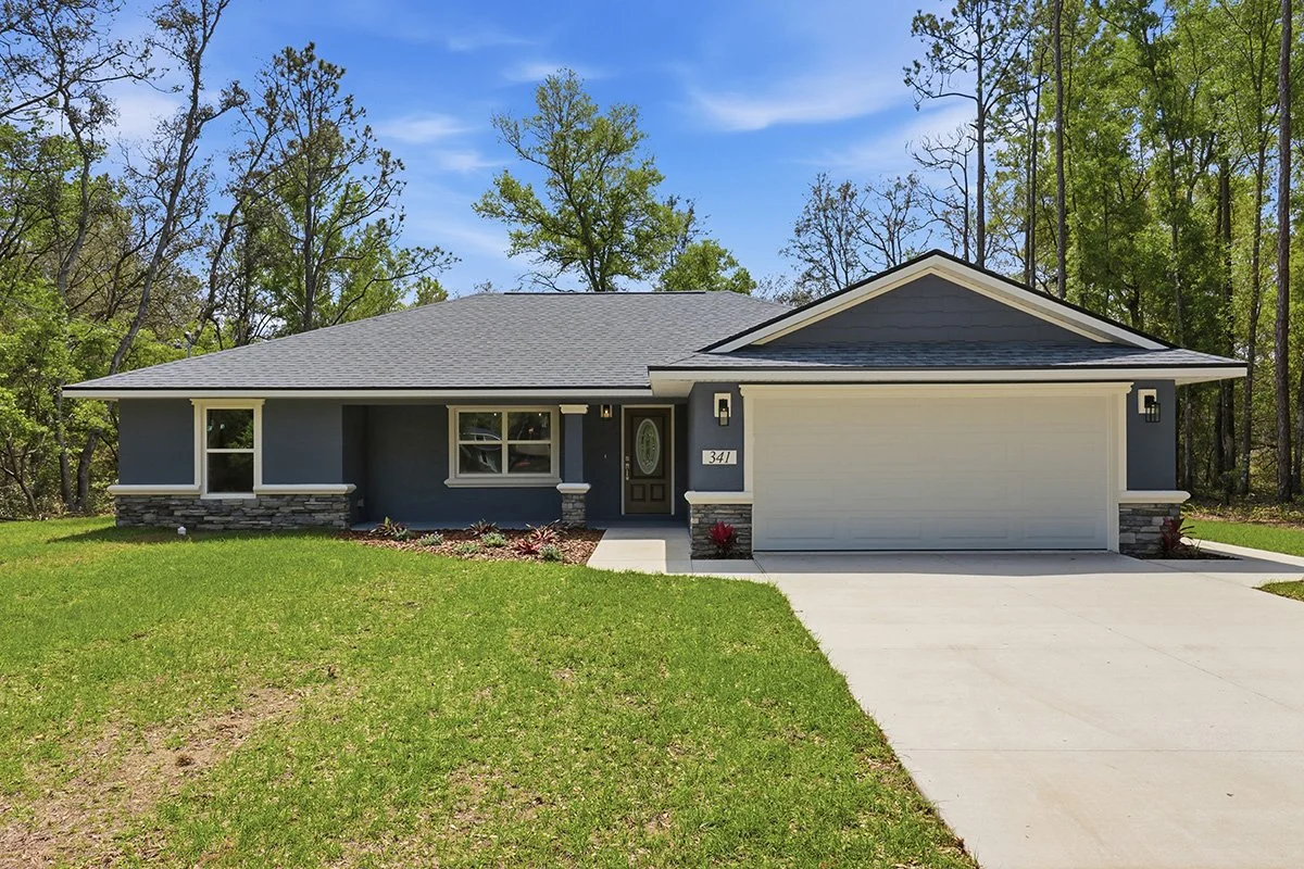  Grayish blue single story Florida home with white banding and white two car garage door. Home has stacked stone accents on front, a wood door, concrete driveway, and blue asphalt shingles. Green grass in foreground and tall trees and blue skies in t