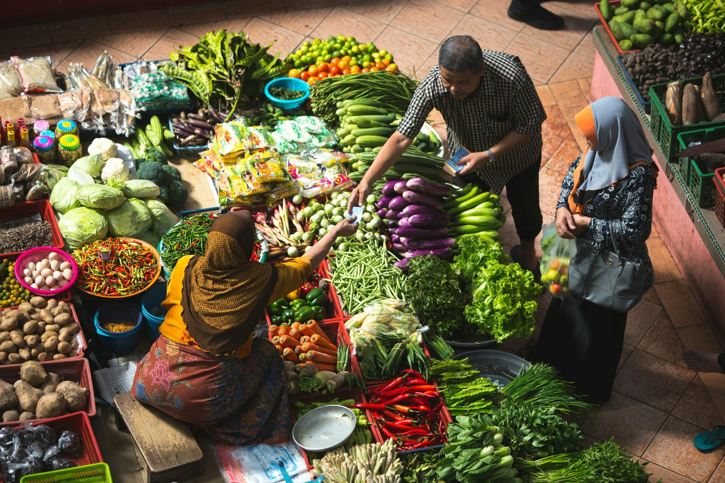 A market stall with various fresh vegetables and fruits, including eggplants, tomatoes, green beans, lettuce, cauliflower, and eggs. A man and woman are shopping, with the woman wearing a headscarf and the man holding a small package.