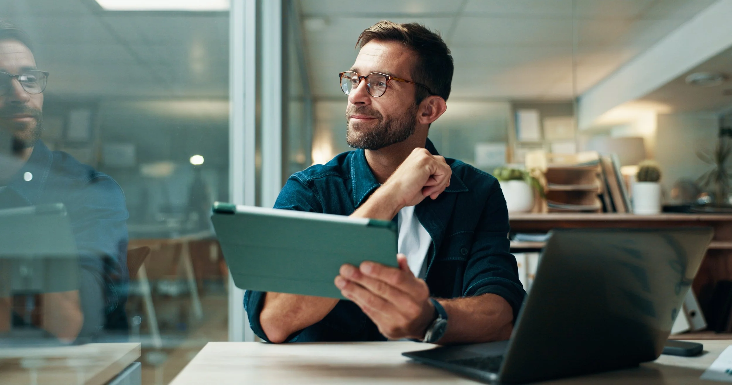 Man with glasses and a beard sitting at a desk, holding a tablet, smiling and looking out of the frame in an office setting.