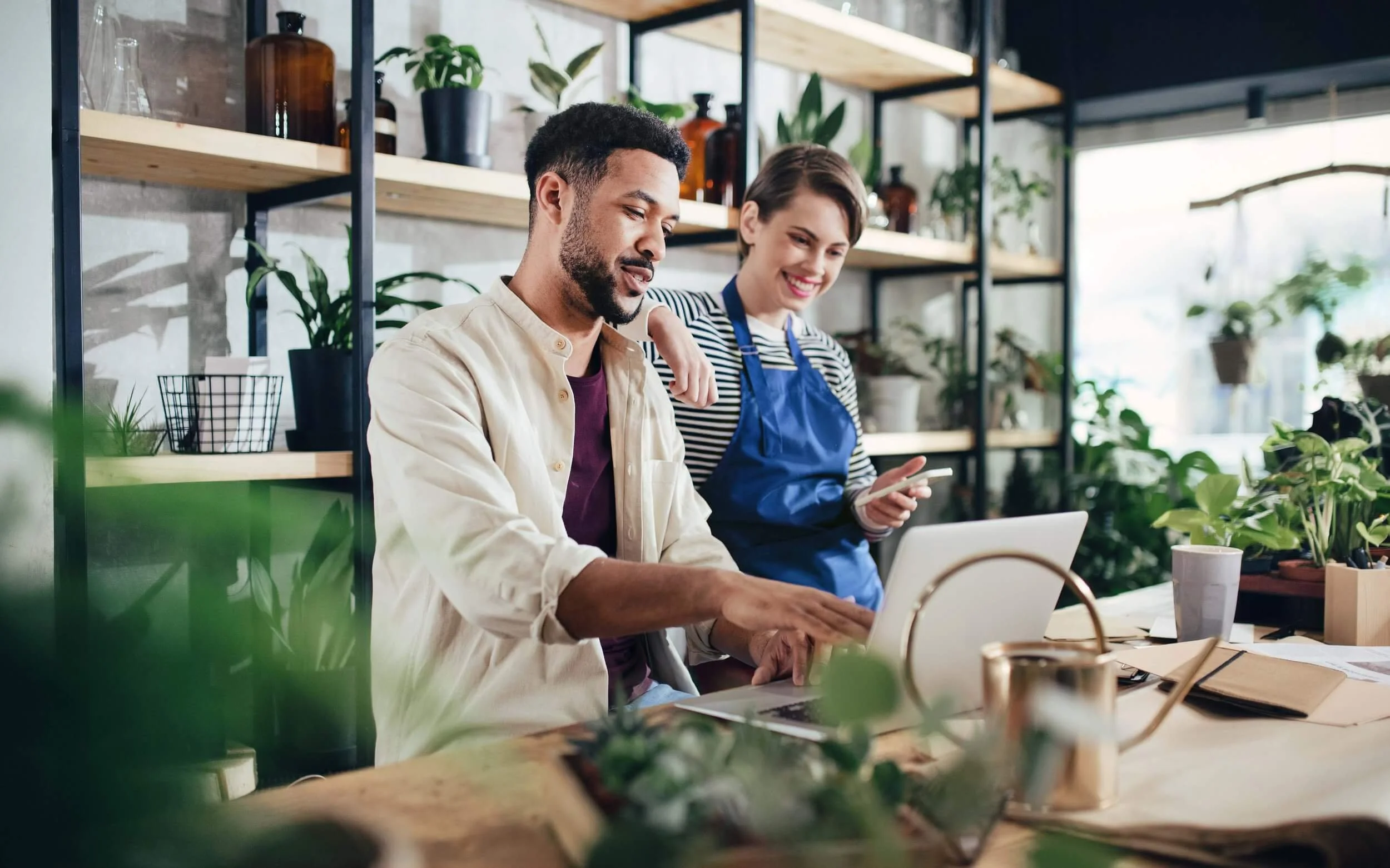 Two people smiling and working together in a plant shop or greenhouse, surrounded by shelves with plants and containers.