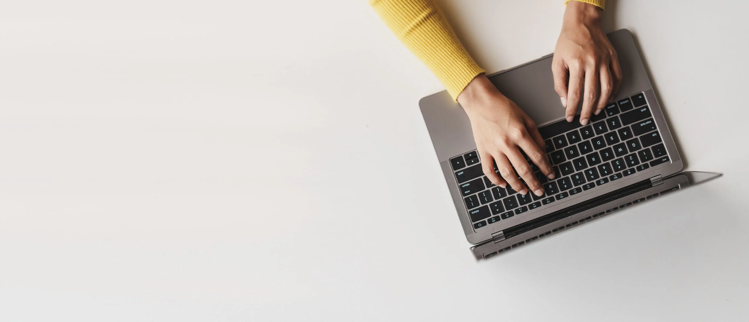 A person wearing a yellow sweater using a silver laptop on a white surface.