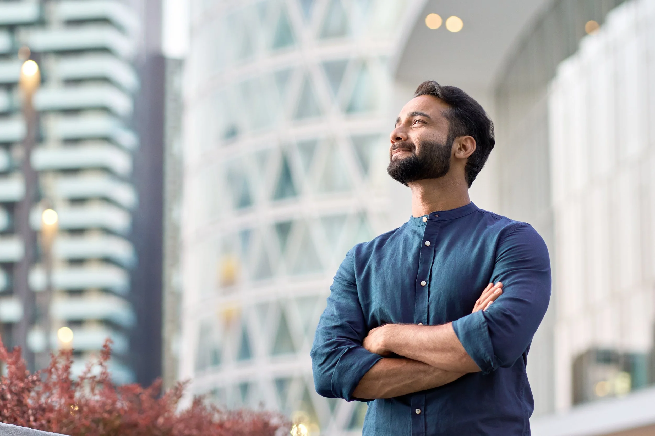 A man with dark hair and a beard, wearing a navy blue shirt, stands outdoors with arms crossed, looking upwards with a smile. Behind him is a modern glass building with reflections and blurred lights.