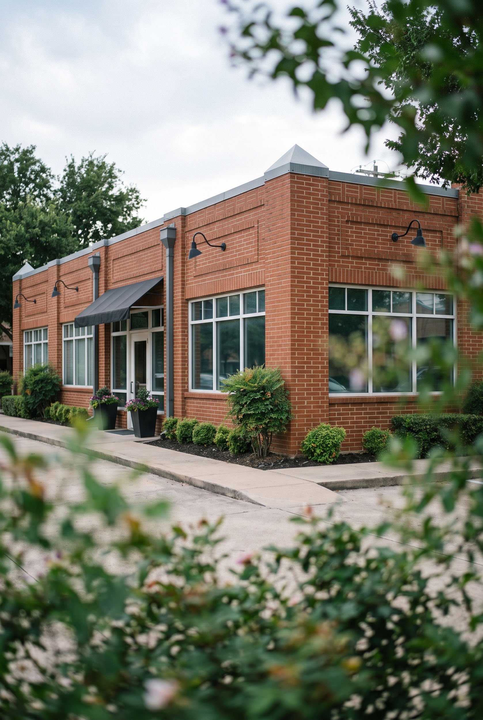 Exterior view of KARPOS therapy practice in Southlake, TX. The building is red brick with large windows, black outdoor wall lights, and surrounded by landscaped bushes and trees.