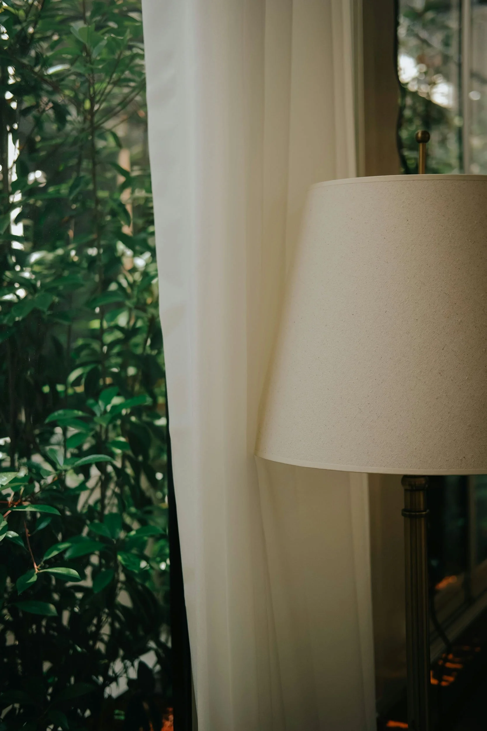 Close-up of a beige lampshade on a floor lamp next to a white curtain, with greenery and a window in the background.