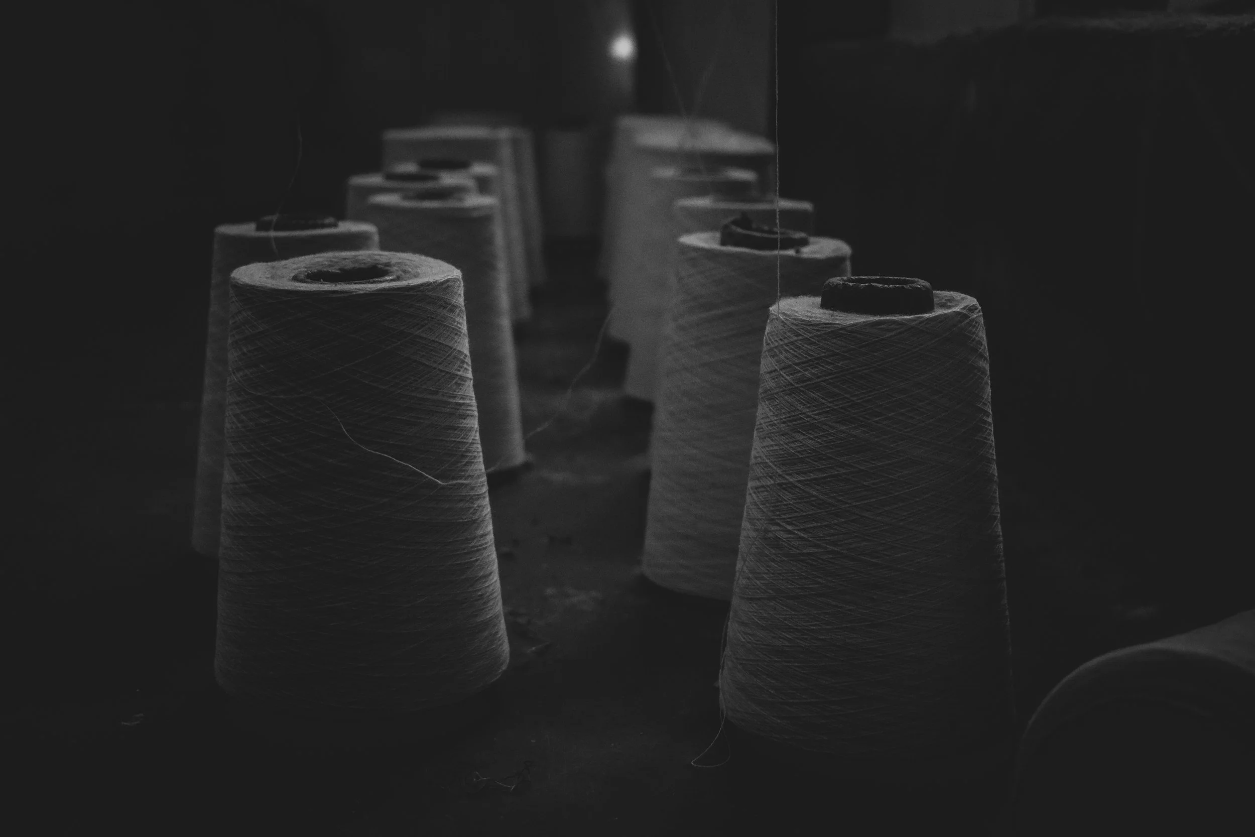 Row of large spools of thread in a factory, illuminated by dim, focused lighting, black and white photograph.
