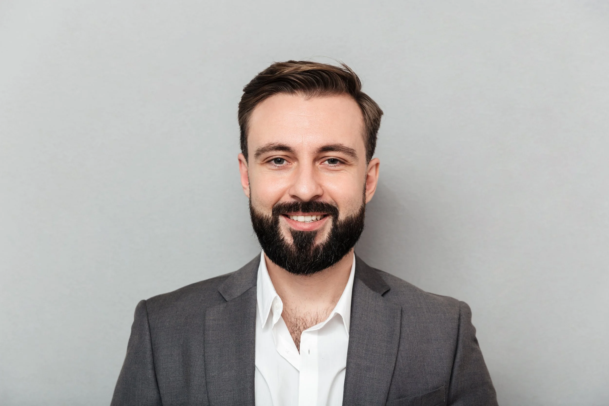Portrait of a smiling man with dark hair and beard wearing a gray suit and white shirt against a light gray background.