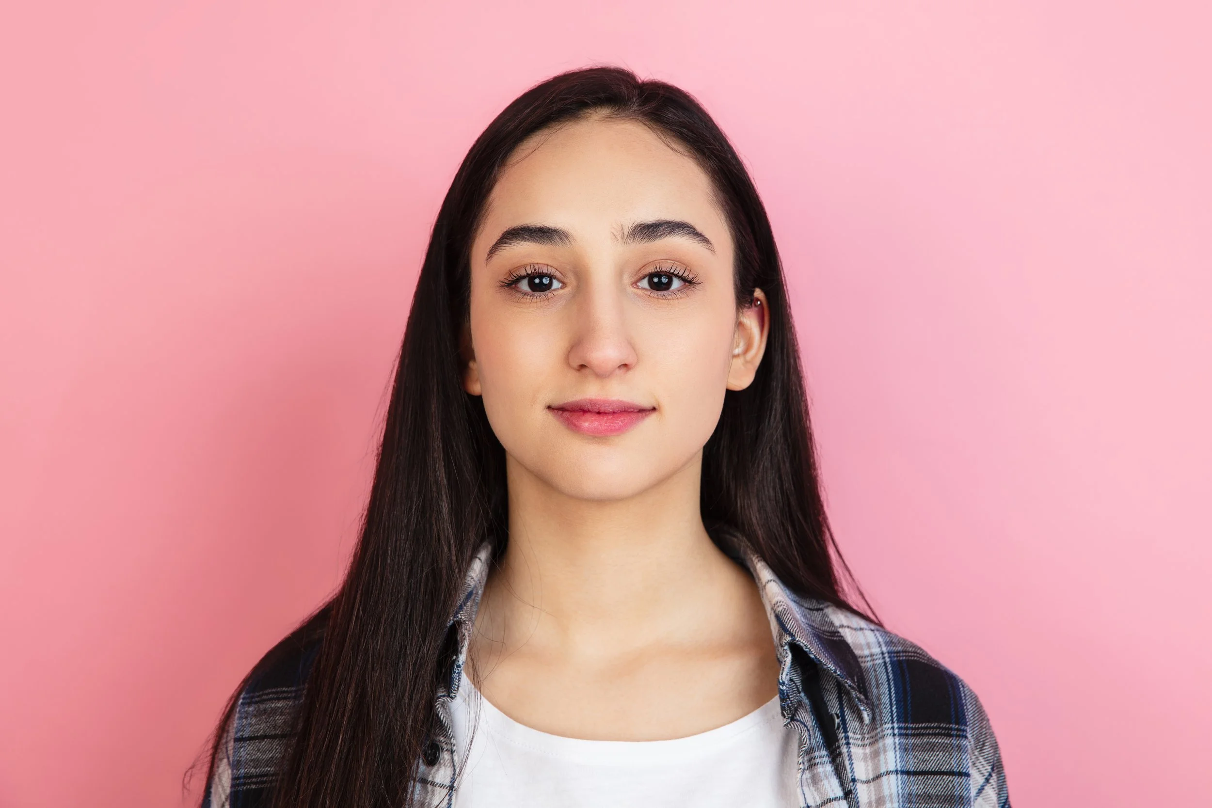 Portrait of a young woman with long dark hair wearing a plaid shirt and white top, smiling softly, against a pink background.