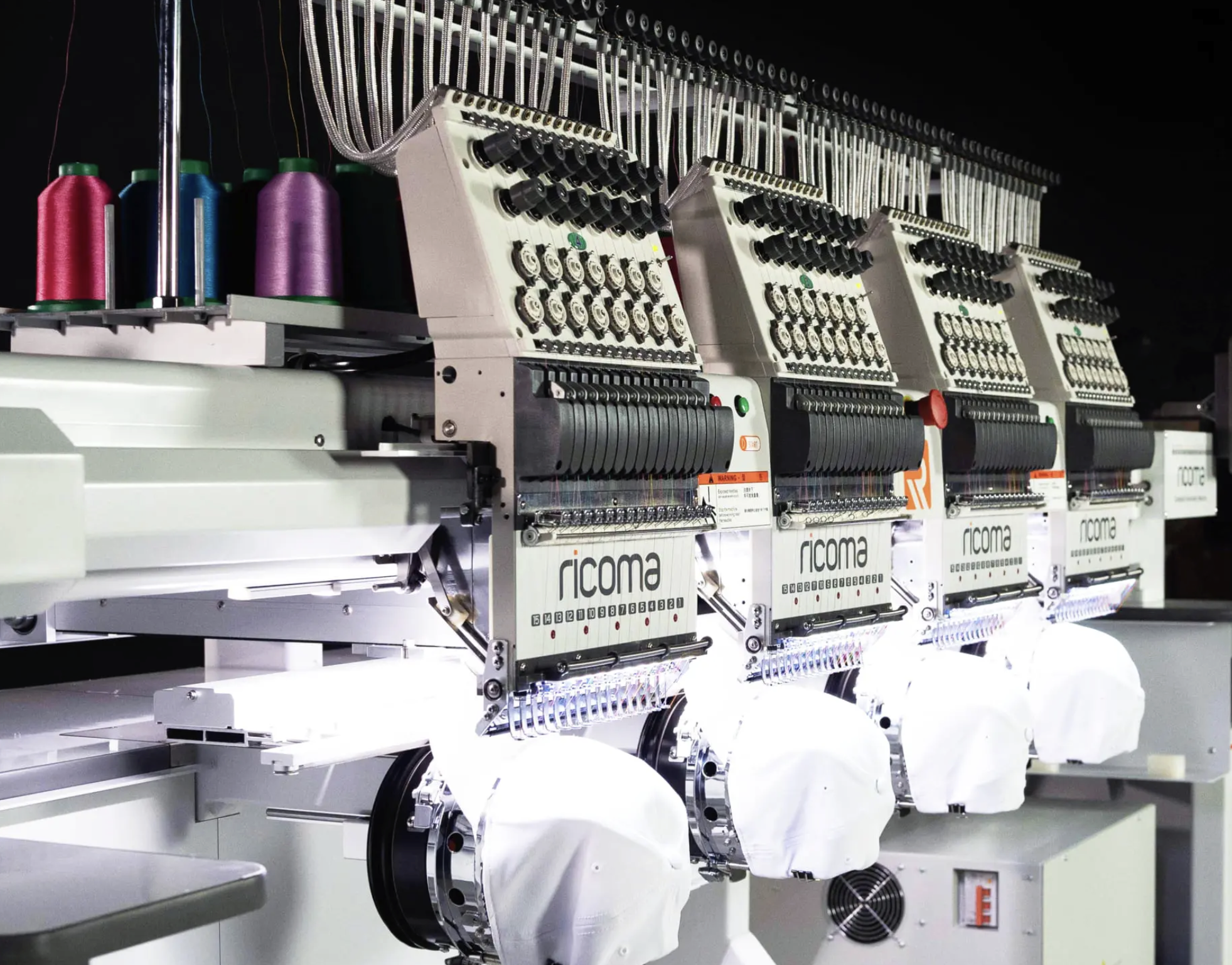 Close-up of multiple industrial embroidery machines with spools of colorful thread on top, working on white fabric.