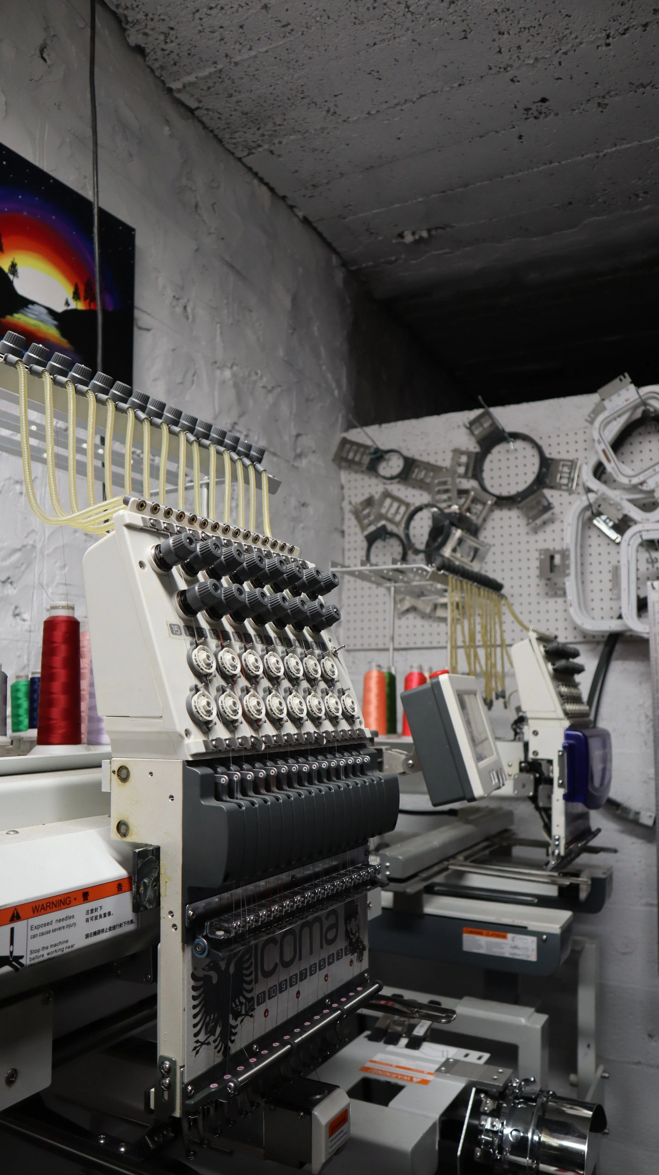 Embroidery sewing machine with spools of thread in a workshop with a textured white wall and a section of artwork showing a sunset with mountains and trees.