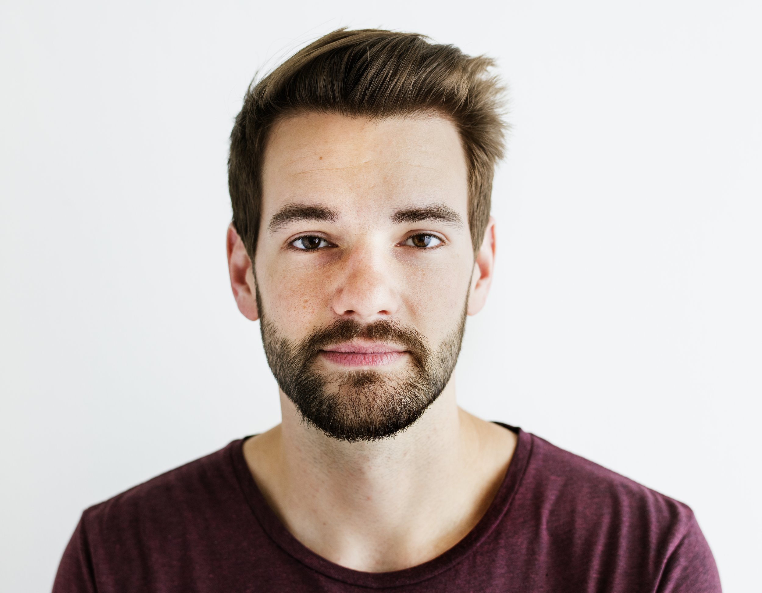 Close-up portrait of a young man with light skin, brown hair, beard, and wearing a maroon shirt against a plain white background.