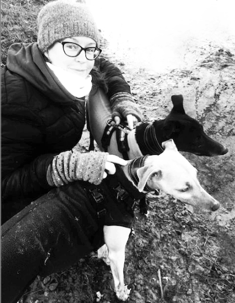Tree Blackmore, a queer teacher for neurodivergent adult English learners, with her whippets in a muddy field. One dog is black and the other pale colour. They are looking to the right and wearing dog fleeces. Tree is wearing a hat and gloves.