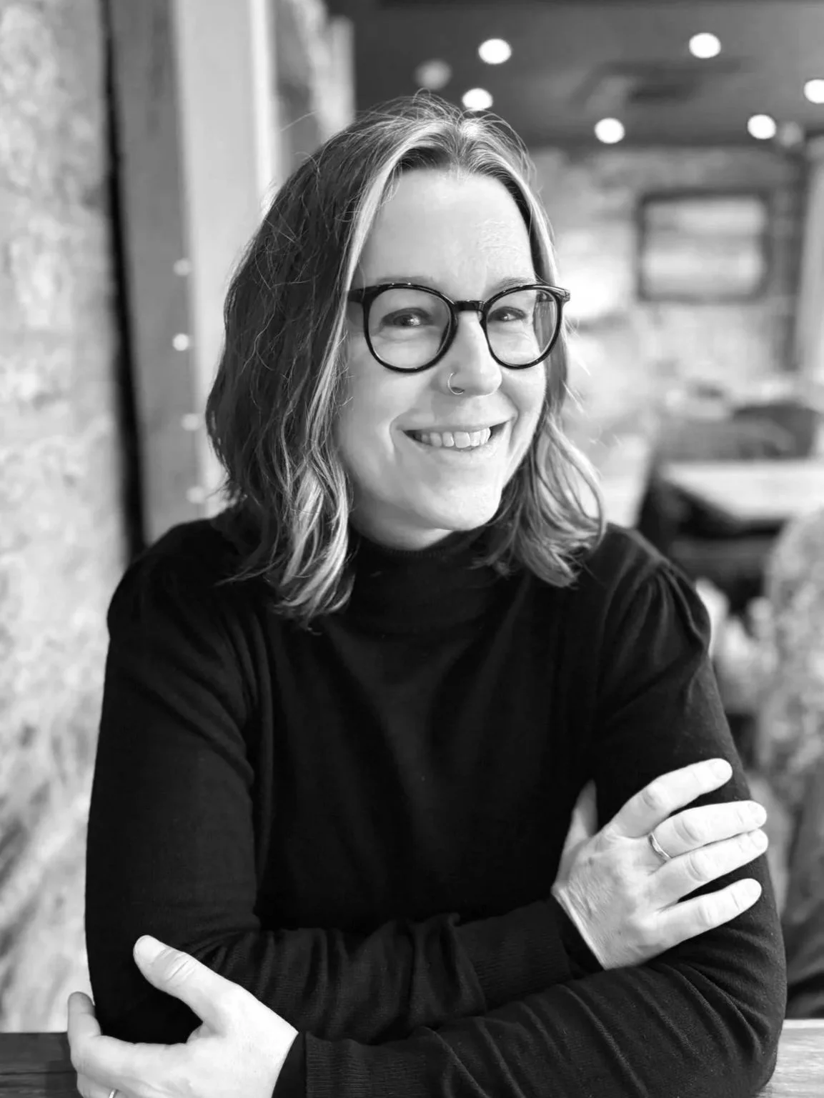 Tree a cis woman with wavy shoulder length hari, glasses and a nose ring is smiling at a cafe table,, arms lightly crossed.