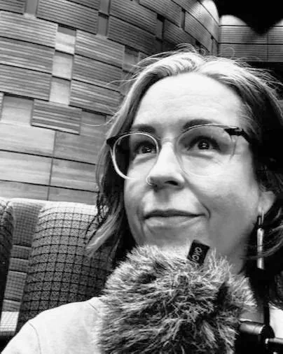 tree blackmore, teacher for LGBTQIA+ English learners, sits in a conference hall with wooden panels. A mic fills the bottom of the photo, which is black and white.