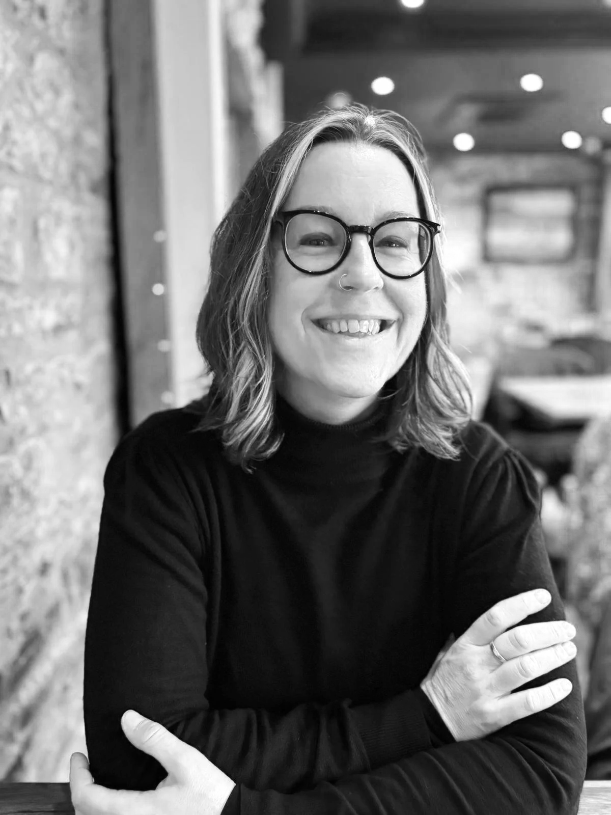 Tree Blackmore, teacher for neurodivergent English students, facing the camera, arms lightly folded. She  has black rimmed glasses and shoulder length hair. the photo is black and white.