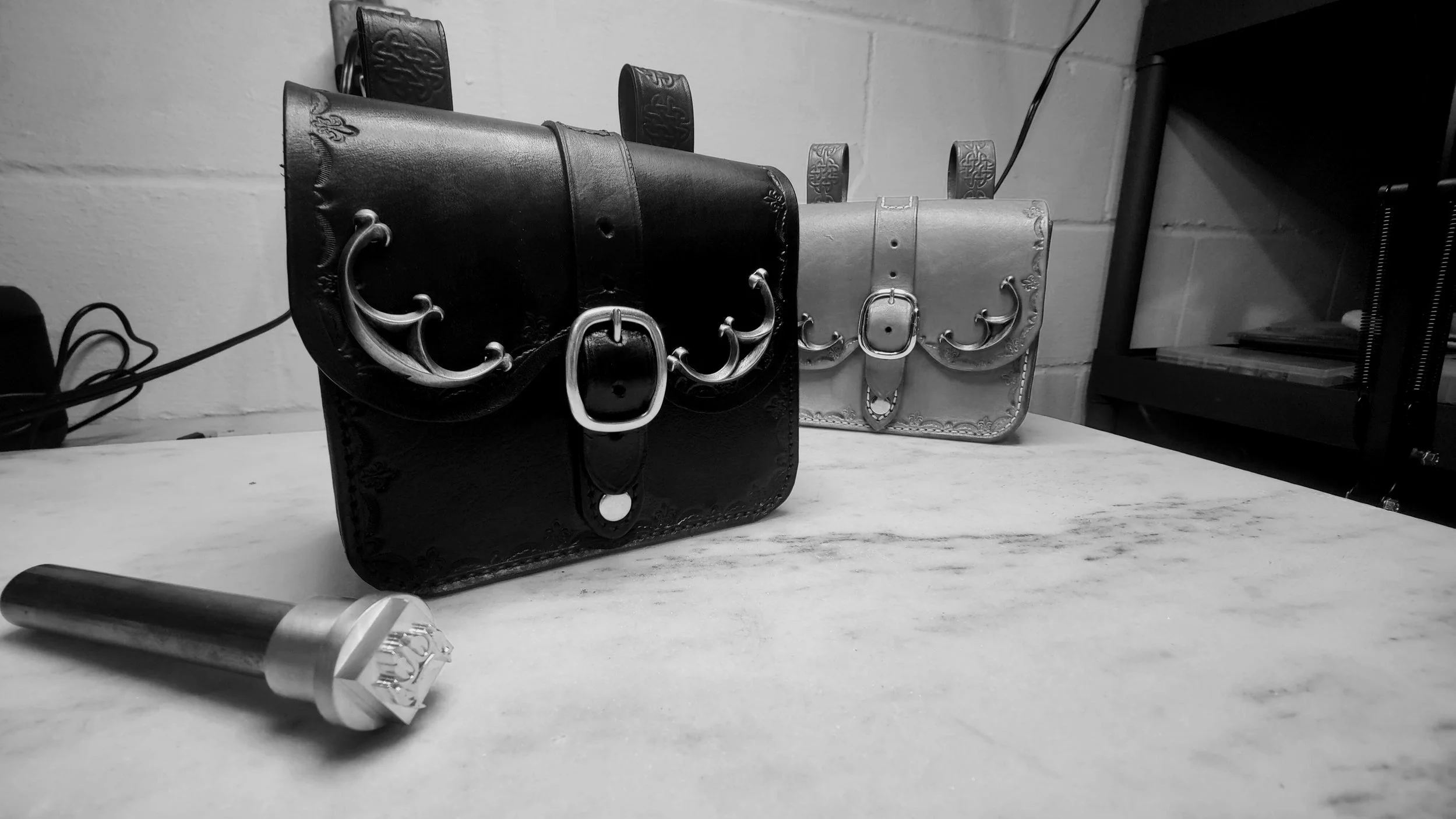 Black and white photo of two decorative leather handbags with metal buckles and floral embossing, placed on a table.
