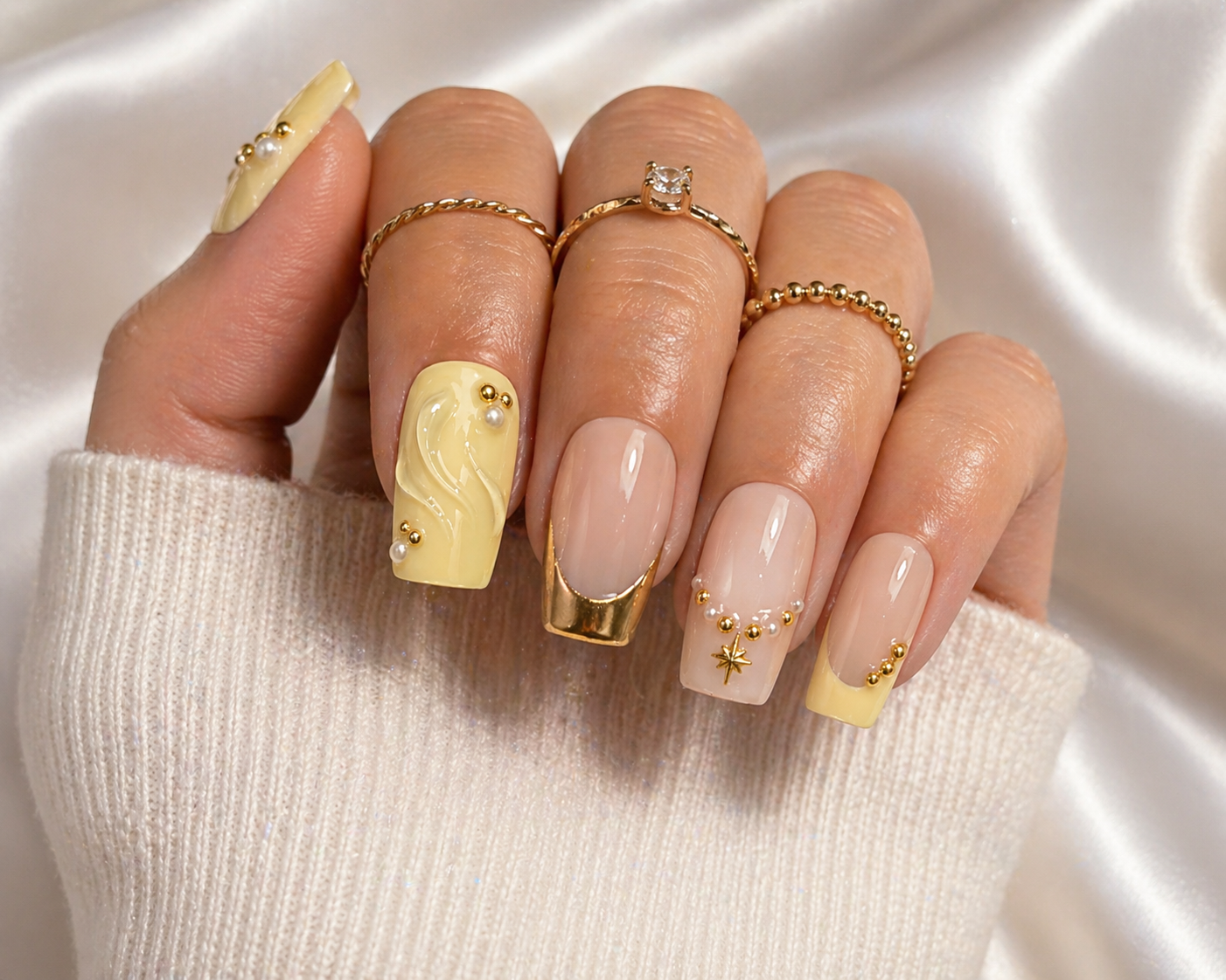 Close-up of a hand with decorated nails and jewelry, wearing a cream-colored sweater. The nails are painted with a light yellow design featuring pearls and golden accents. The person is wearing multiple rings including a twisted band, a solitaire ring, and a beaded band.