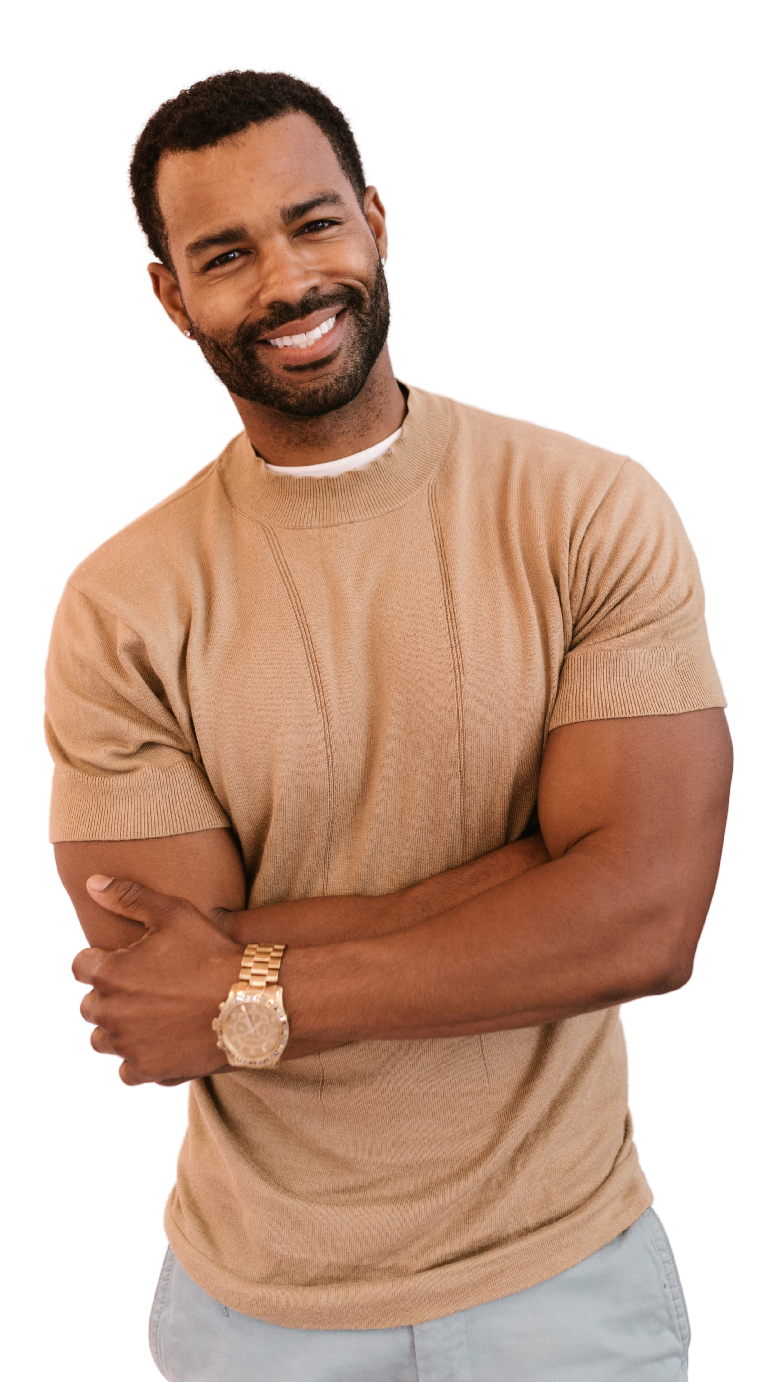 A smiling man with a beard and short curly hair, wearing a beige t-shirt, gold watch, and light-colored jeans, crossing his arms.