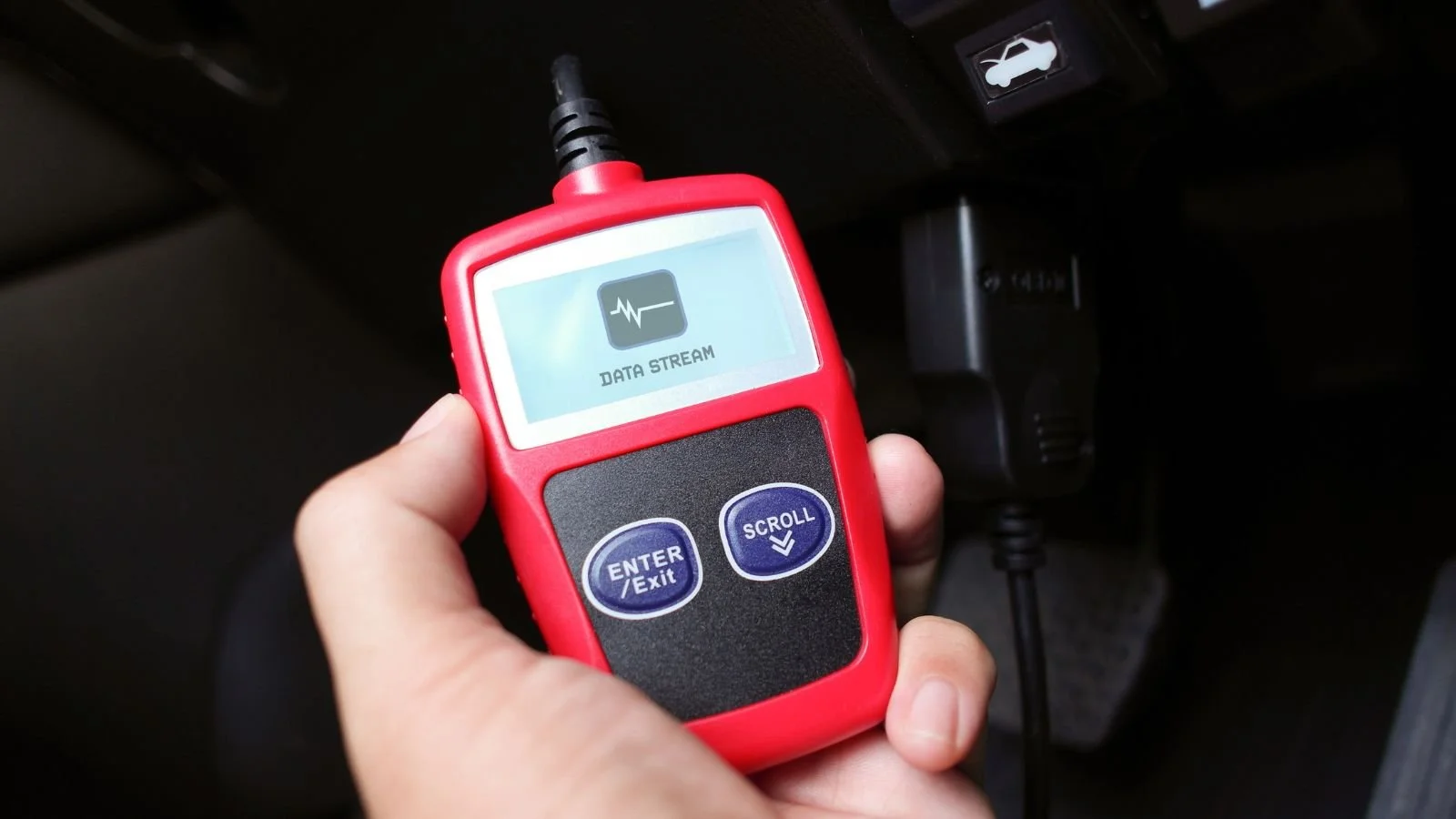 Handheld diagnostic tool with a screen labeled 'Data Stream' and two buttons labeled 'Enter Exit' and 'Scroll', held near a car's dashboard and power outlet.
