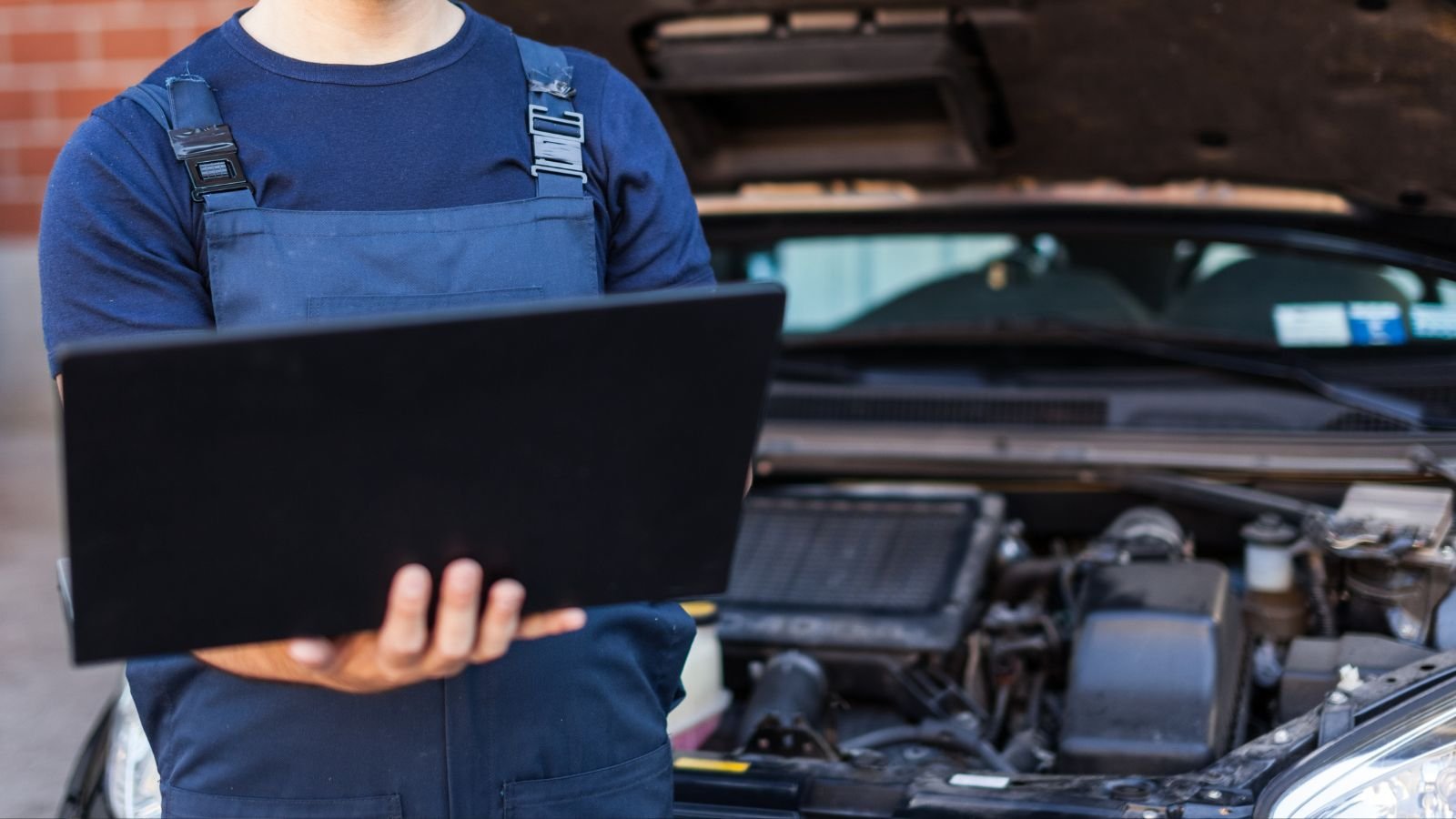 A mechanic wearing a blue uniform holds a laptop in front of an open car hood, inspecting the engine.