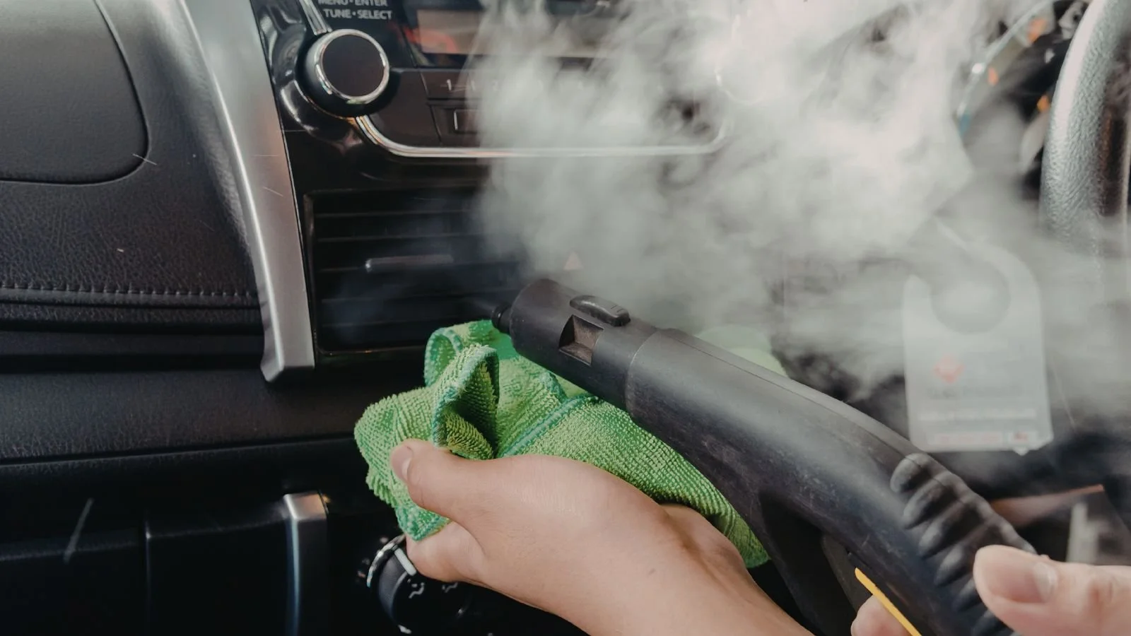Person using a steam cleaner with a green towel to clean the air vents of a car interior.