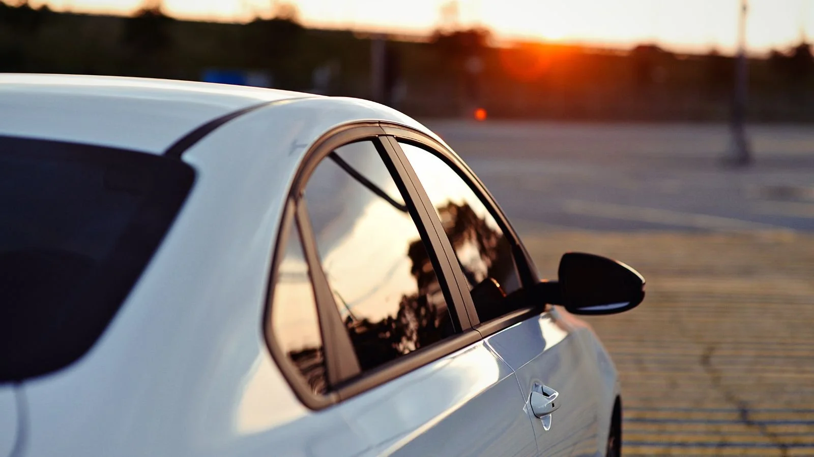 Close-up of a white car parked in an empty lot during sunset, with reflections of trees in the windows.