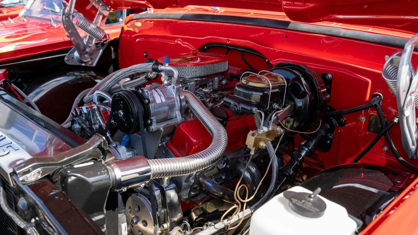 Close-up of a vintage car engine bay with shiny metal components, red-painted inner fenders, and various hoses and wires.