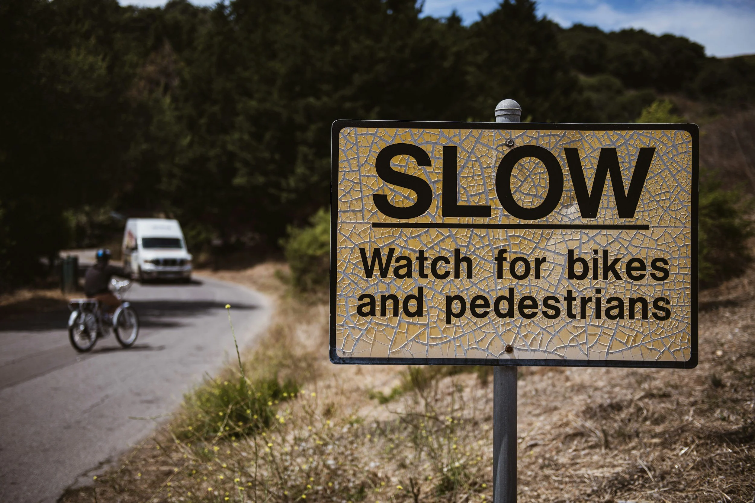 A road sign that says 'SLOW' and 'Watch for bikes and pedestrians', with a background of a winding road, a cyclist, and an approaching vehicle.