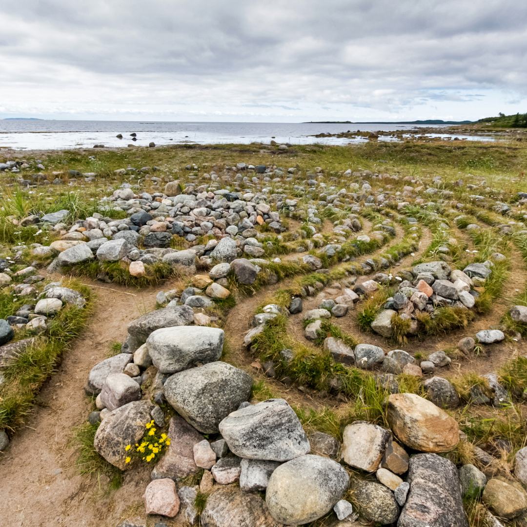 A rocky coastal landscape with a winding stone path leading to the beach, grassy areas, and cloudy sky over the ocean in the background.