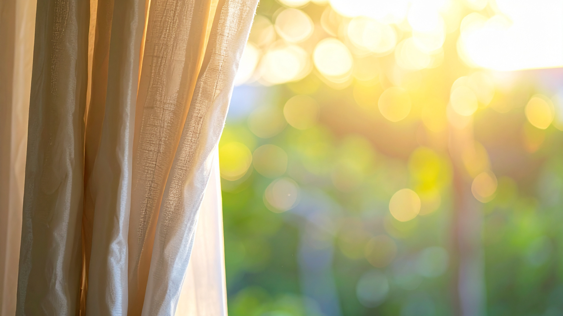 Close-up of beige curtains with sunlight streaming through a window, creating a bokeh effect in the background.